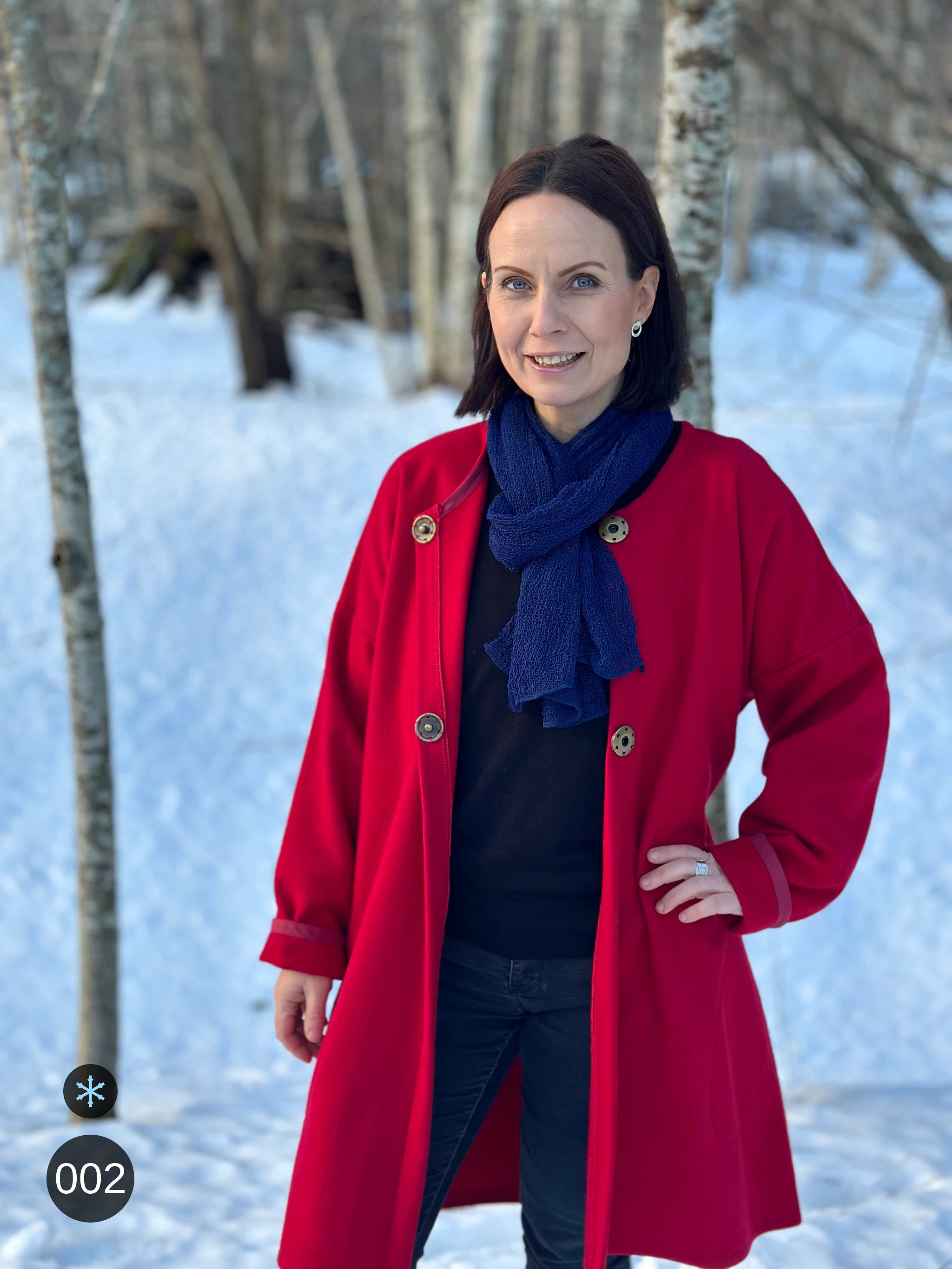 Kvinna bär Almira Sjal i färg 002 Marinblå, en kall färg i djup marinblå, lång finstickad unisex-sjal som faller mjukt nedför framsidan, fotad över en röd kappa i snöig björkskog. Ulla Jacobsson Kläder.