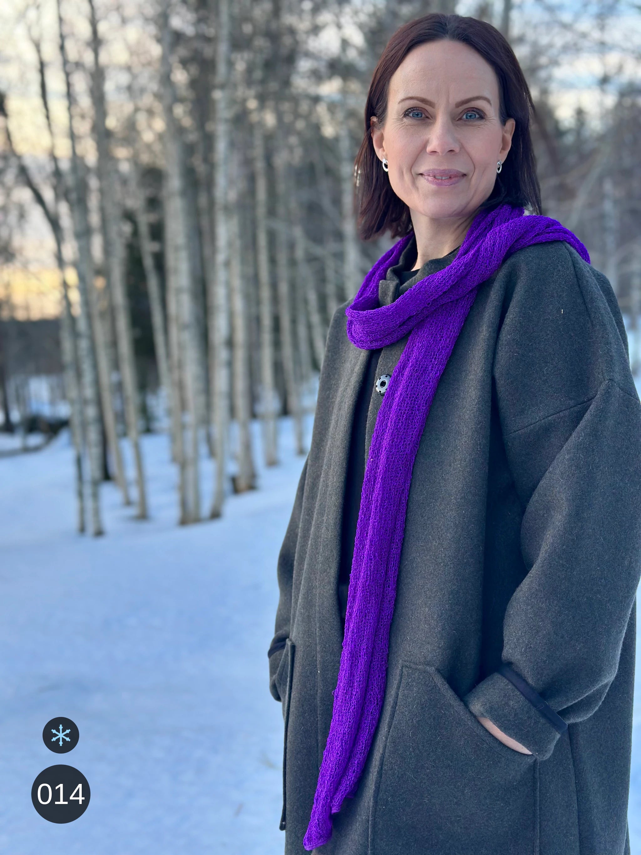 Kvinna bär Almira Sjal i färg 014 Mörklila, en kall färg i klar violett lila, lång finstickad unisex-sjal som faller mjukt nedför framsidan, fotad över en grå kappa i snöig björkskog. Ulla Jacobsson Kläder.