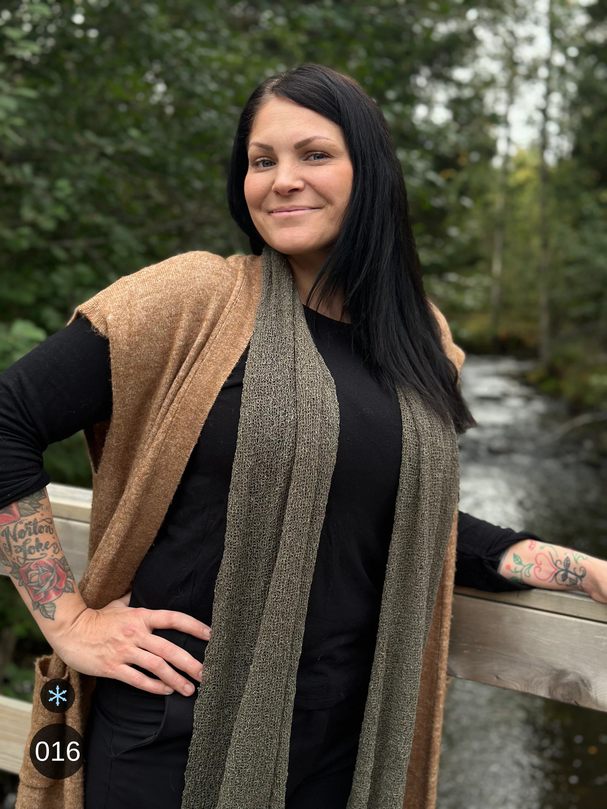 Kvinna bär Almira Sjal i färg 016 Lavgrön, en kall färg i dämpad gråbrun grönton som lav på sten, lång finstickad unisex-sjal som faller mjukt nedför framsidan, fotad vid ett trästaket intill ån. Ulla Jacobsson Kläder.