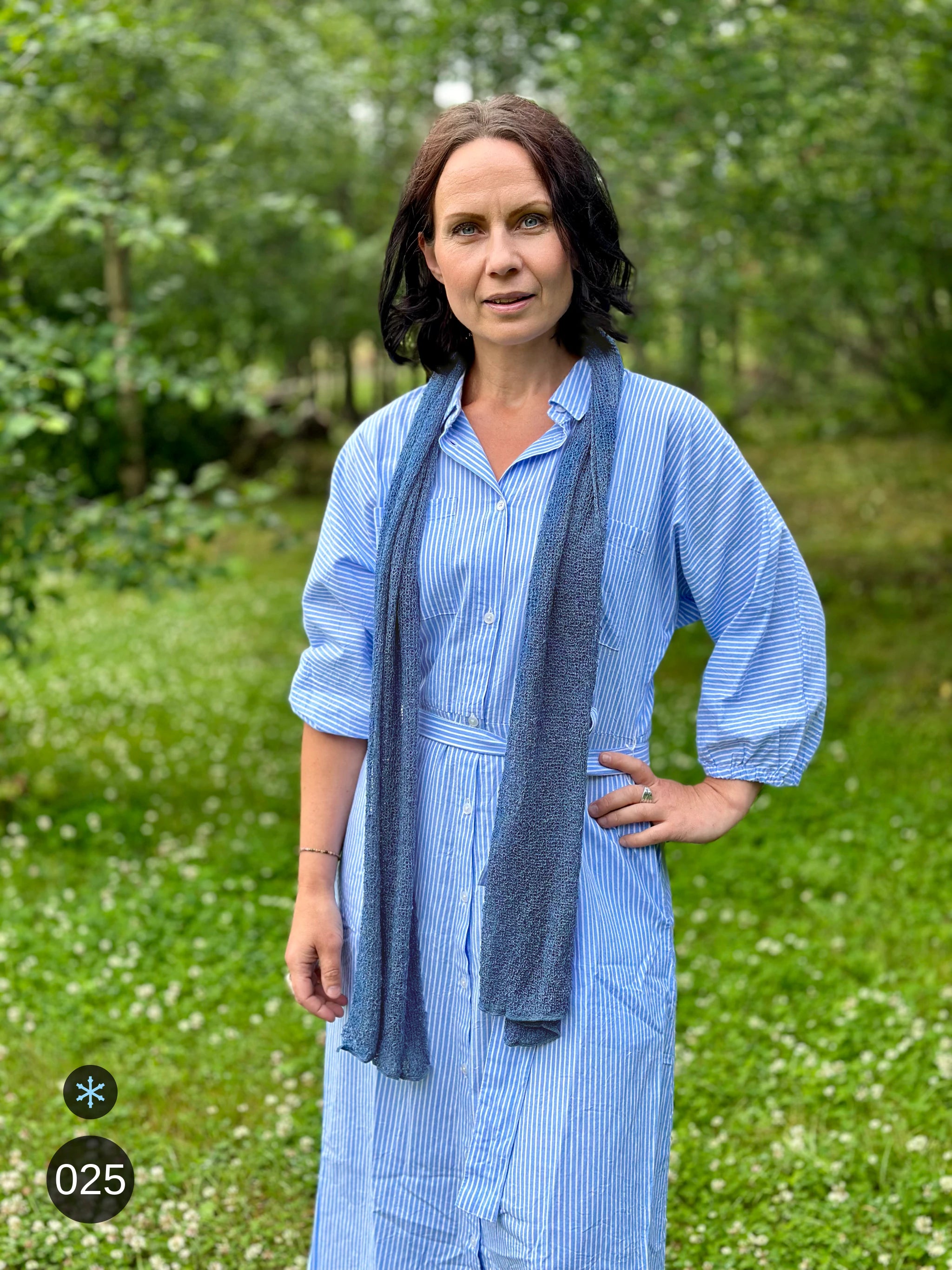 Kvinna bär Almira Sjal i färg 025 Jeansblå, en kall färg i dämpad denimblå, lång finstickad unisex-sjal som faller mjukt nedför framsidan, fotad till en ljusblå linneklänning i sommargrönska. Ulla Jacobsson Kläder.