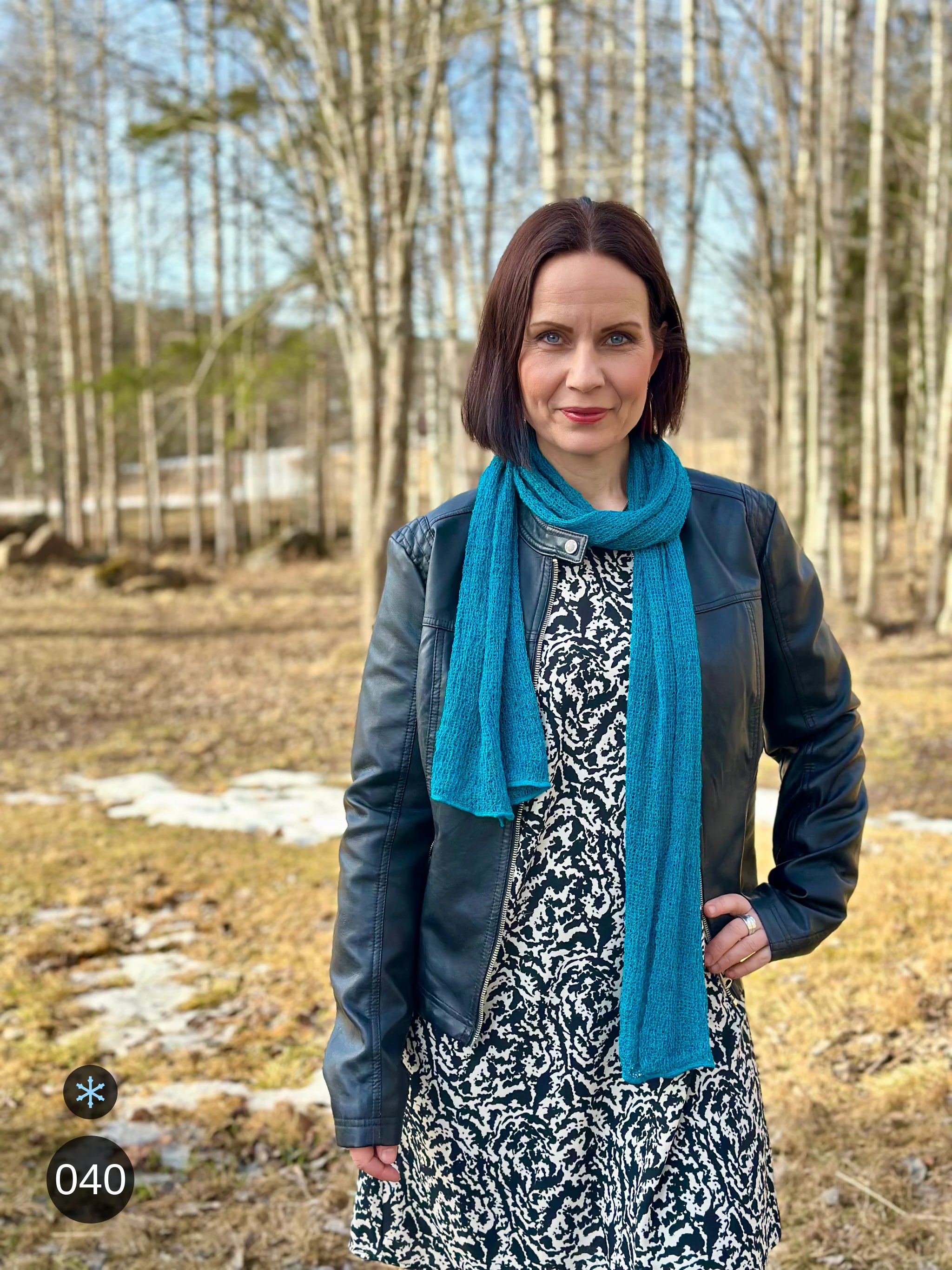 Kvinna bär Almira Sjal i färg 040 Petrol, en kall färg i djup petrolblå, lång finstickad unisex-sjal som faller mjukt nedför framsidan, fotad till en mönstrad klänning och skinnjacka en tidig vårdag. Ulla Jacobsson Kläder.