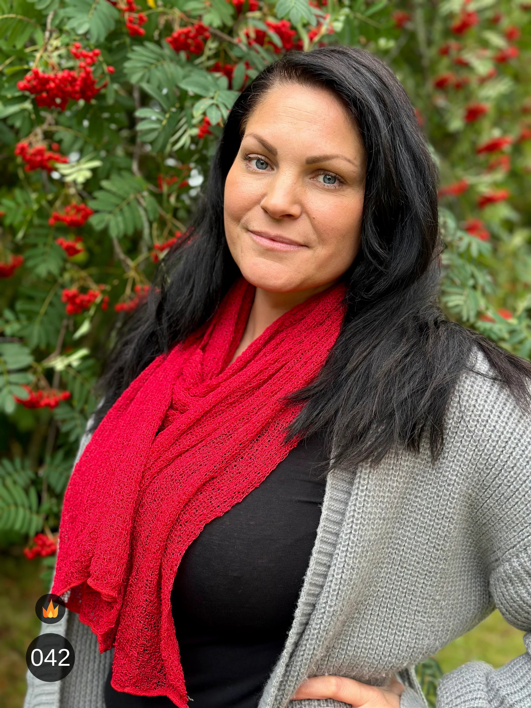 Kvinna bär Almira Sjal i färg 042 Röd, en varm färg i klar röd, lång finstickad unisex-sjal som faller mjukt nedför framsidan, fotad framför en rönn full av röda bär. Ulla Jacobsson Kläder.
