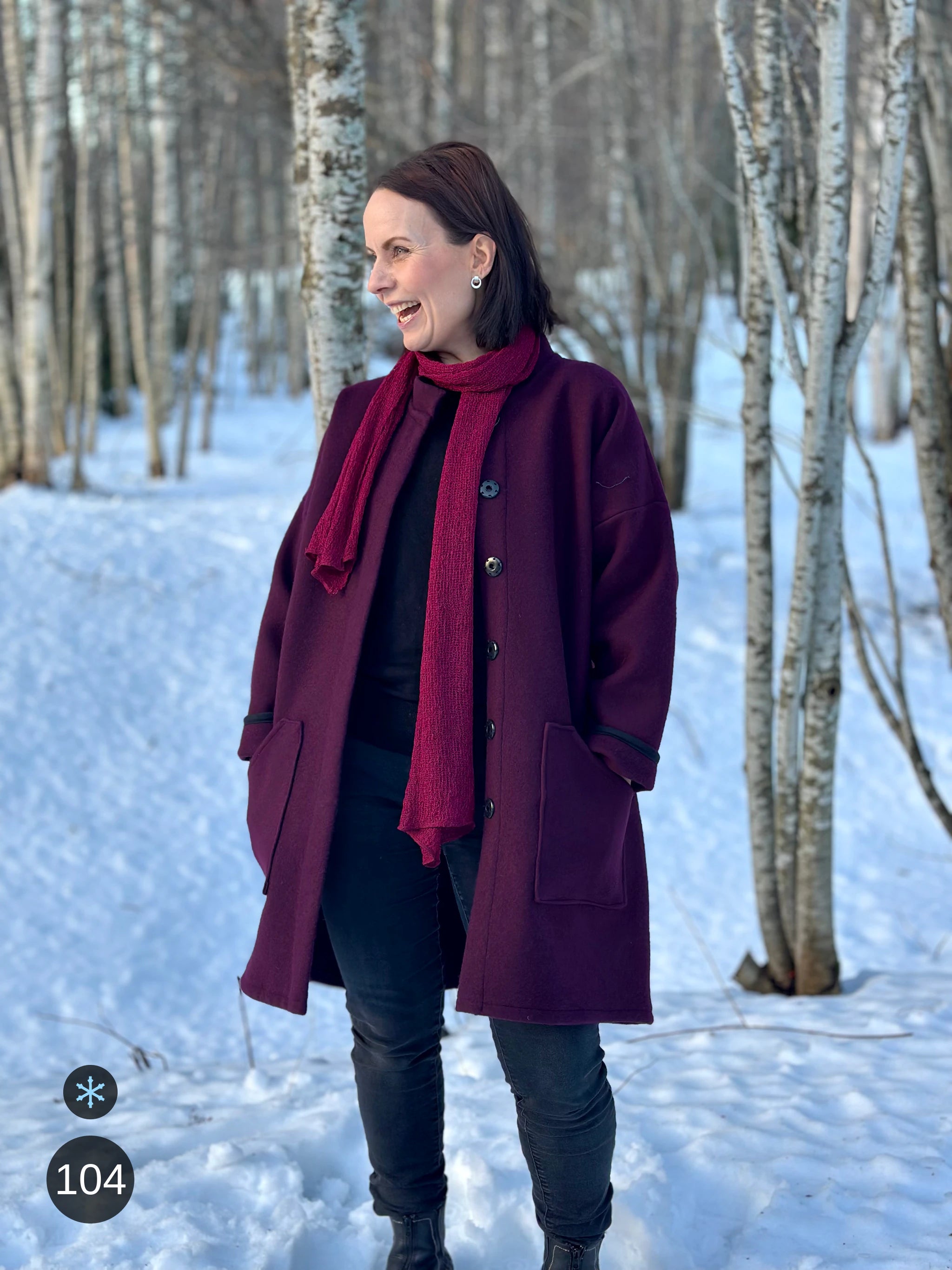 Kvinna bär Almira Sjal i färg 104 Fuchsia, en kall färg i djup fuchsia, lång finstickad unisex-sjal som faller mjukt nedför framsidan, fotad över en vinröd kappa i snöig björkskog. Ulla Jacobsson Kläder.