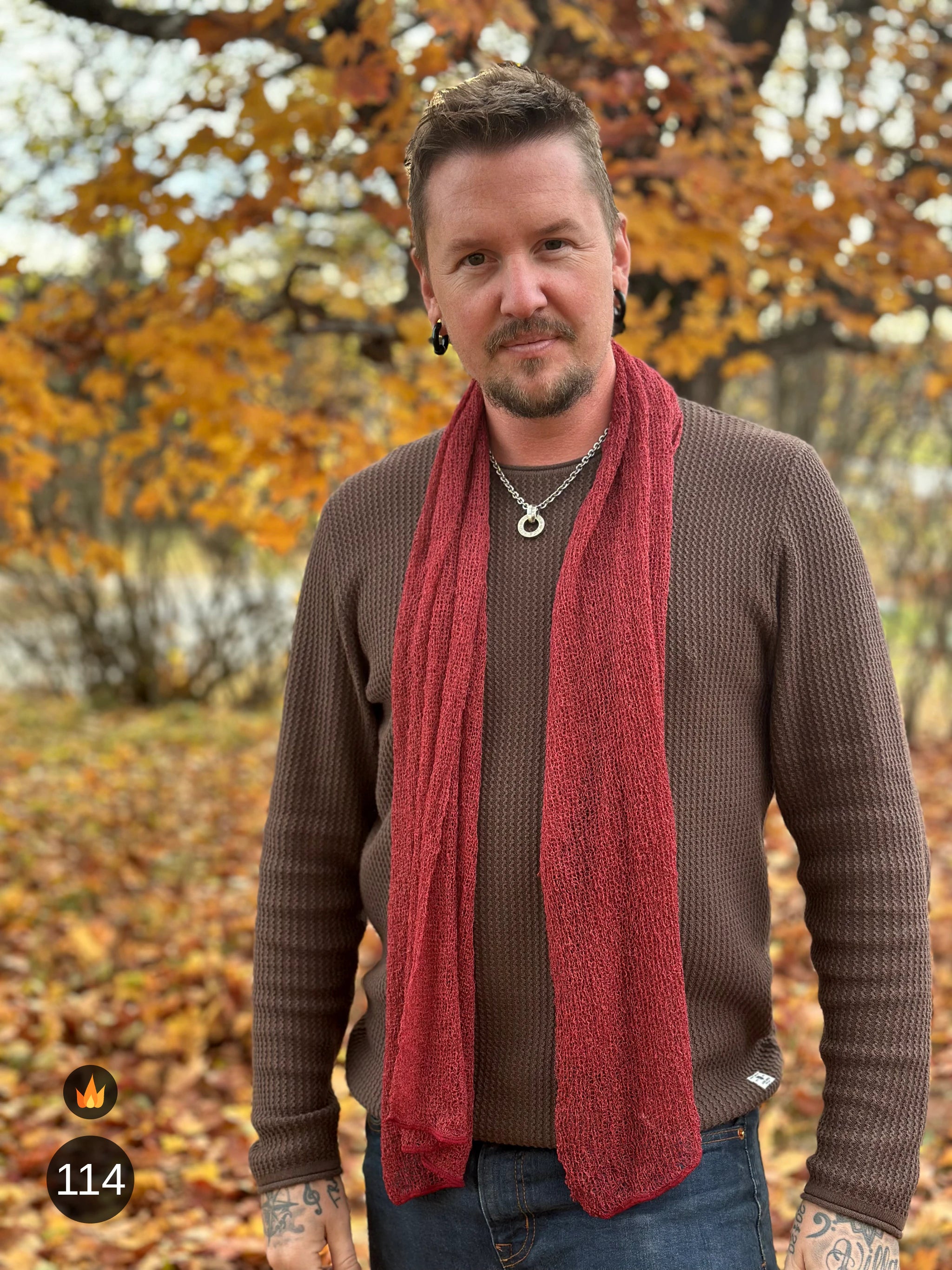Man bär Almira Sjal i färg 114 Maroon, en varm färg i dämpad maroon med tegelton, lång finstickad unisex-sjal som faller mjukt nedför framsidan, fotad bland höstlöv en höstdag. Ulla Jacobsson Kläder.