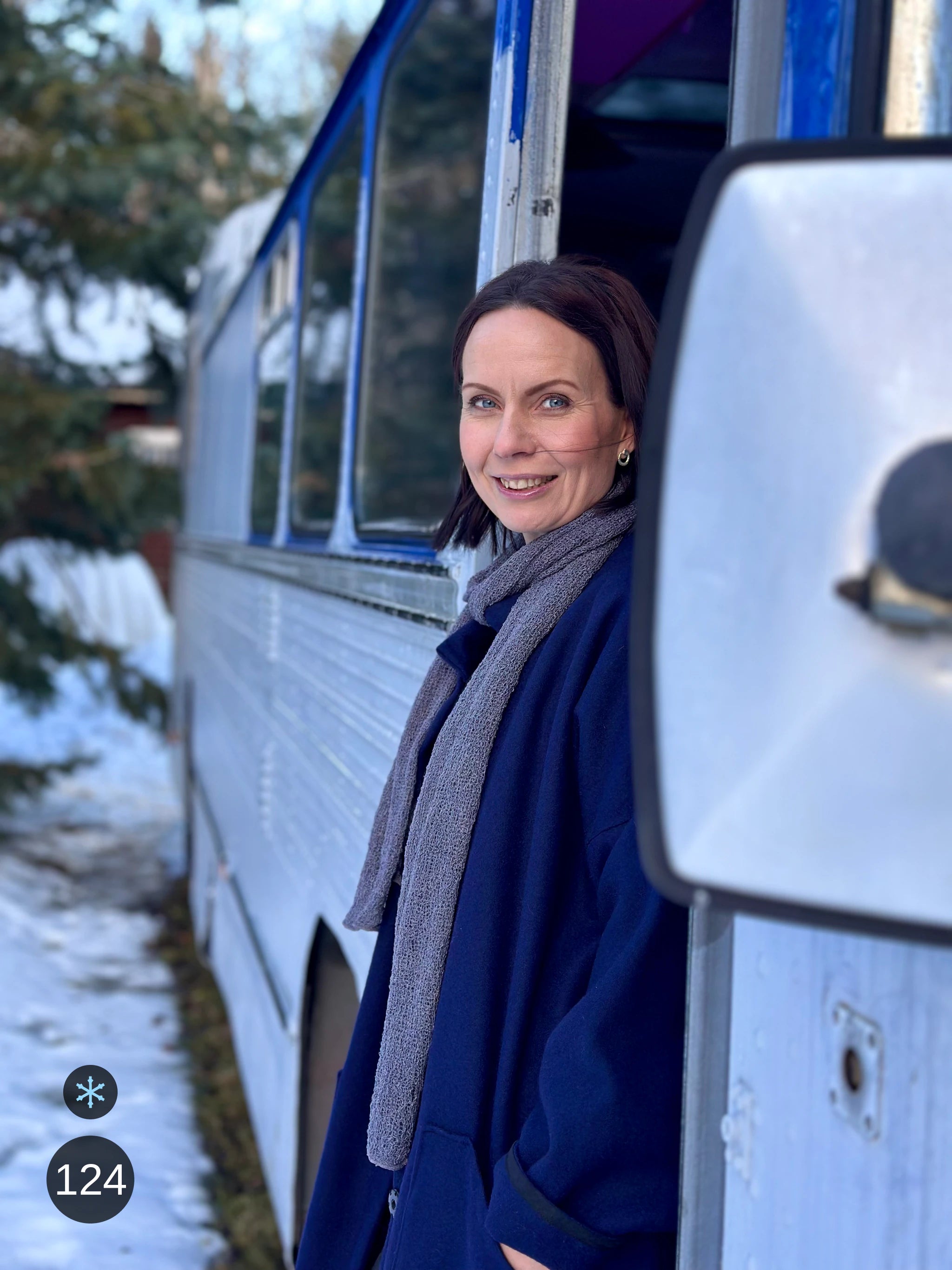 Kvinna bär Almira Sjal i färg 124 Grålila, en kall färg i dämpad grålila, lång finstickad unisex-sjal som faller mjukt nedför framsidan, fotad vid en vit buss. Ulla Jacobsson Kläder.