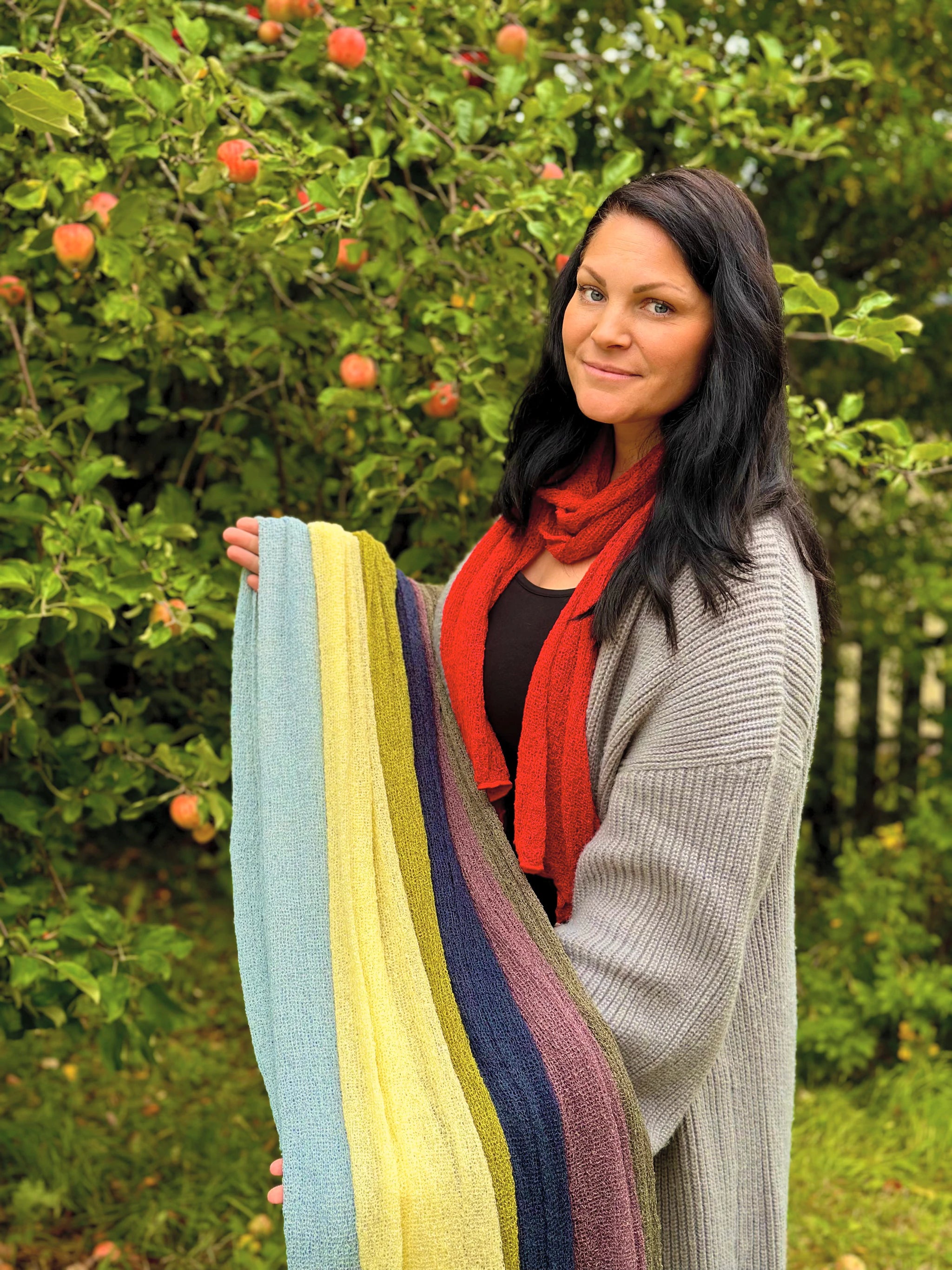 Kvinna bär Almira Sjal i rött och håller upp en bunt sjalar i flera färger, långa finstickade unisex-sjalar, fotad under ett äppelträd en septemberdag. Ulla Jacobsson Kläder.