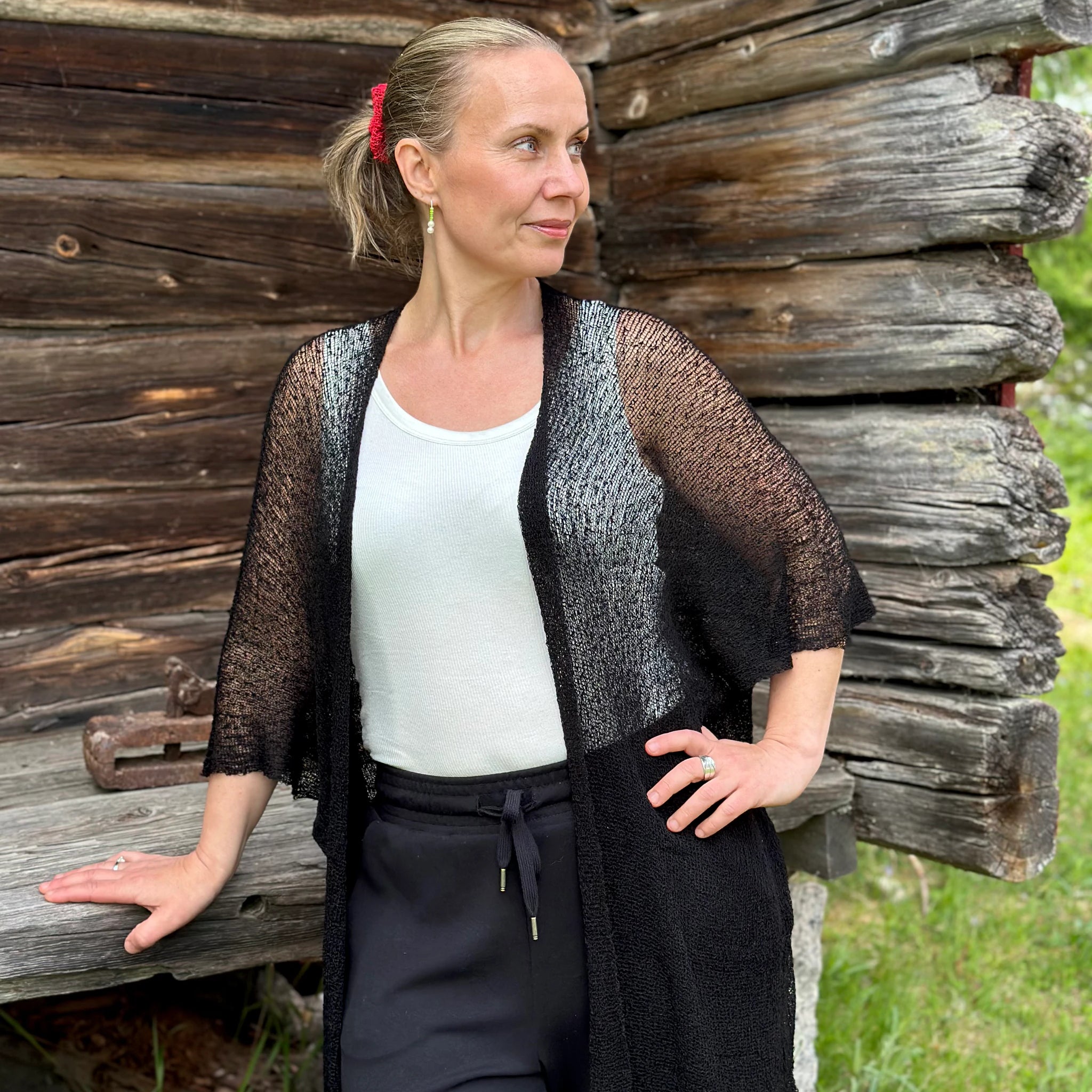 Kvinna bär Biasa Kofta i färg 001 Svart, en neutral djupsvart med matt yta, lång finstickad oversized kofta buren öppen, fotad framför en gammal timmervägg i sommargrönska. Ulla Jacobsson Kläder.
