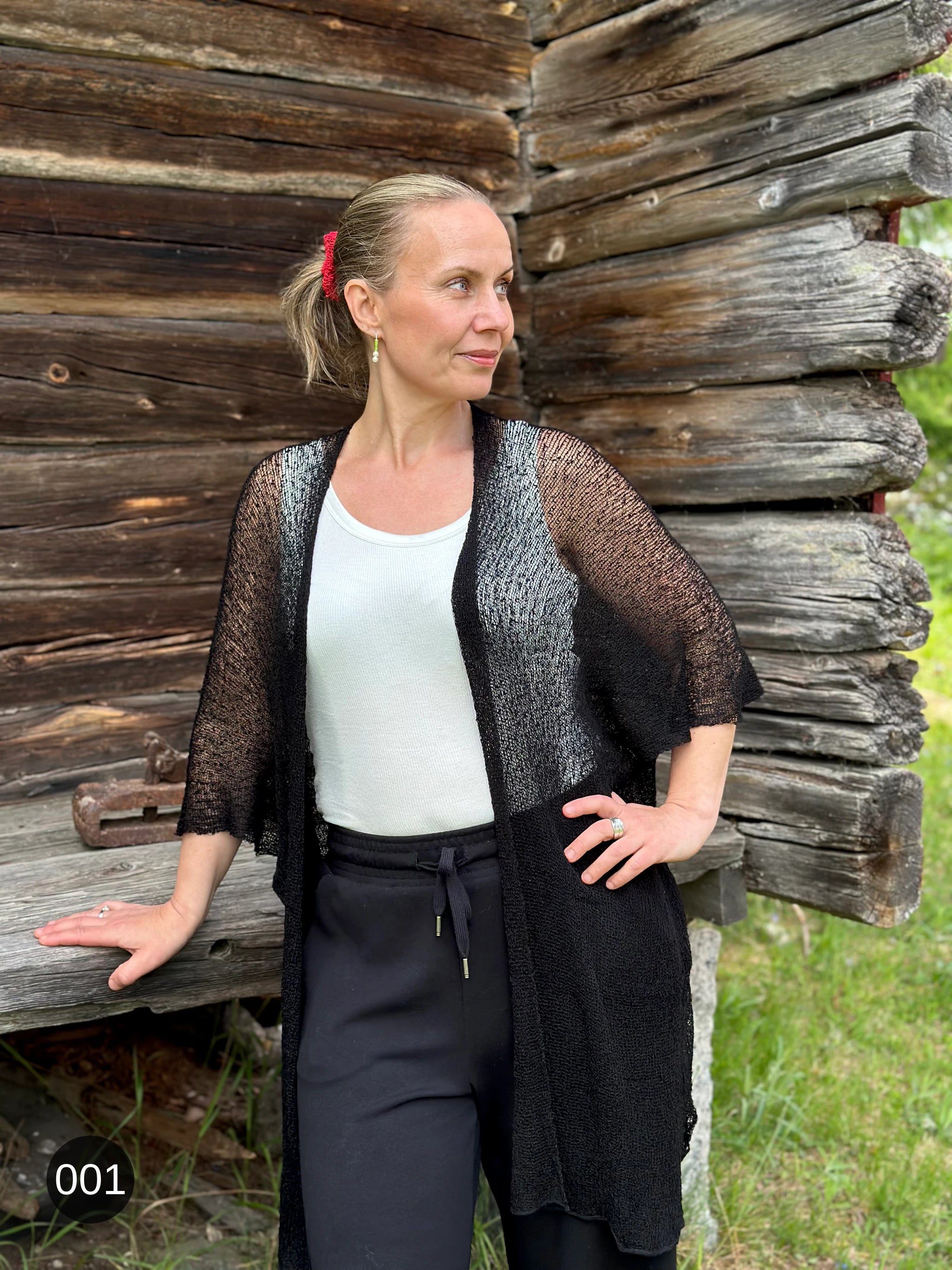 Kvinna bär Biasa Kofta i färg 001 Svart, en neutral djupsvart med matt yta, lång finstickad oversized kofta buren öppen, fotad framför en gammal timmervägg i sommargrönska. Ulla Jacobsson Kläder.