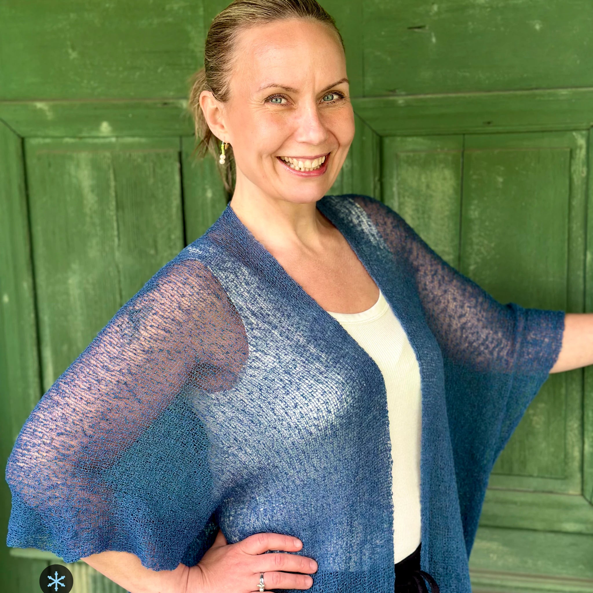 Kvinna bär Biasa Kofta i färg 025 Jeansblå, en kall färg i dämpad denimblå, lång finstickad oversized kofta buren öppen, fotad framför en grön trädörr i sommarsol. Ulla Jacobsson Kläder.