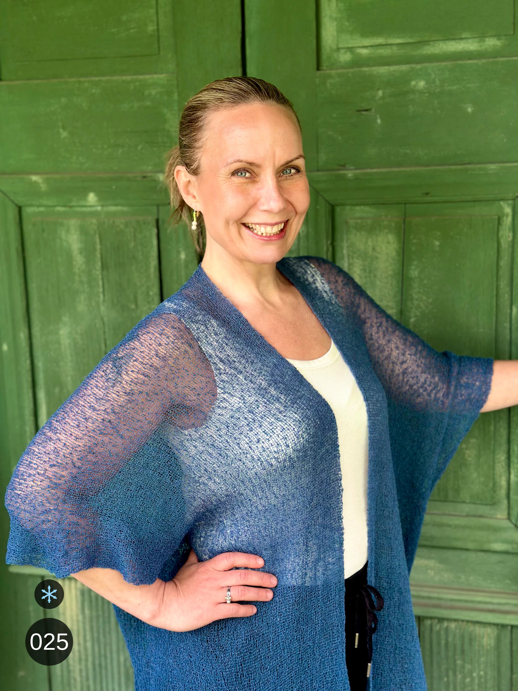 Kvinna bär Biasa Kofta i färg 025 Jeansblå, en kall färg i dämpad denimblå, lång finstickad oversized kofta buren öppen, fotad framför en grön trädörr i sommarsol. Ulla Jacobsson Kläder.