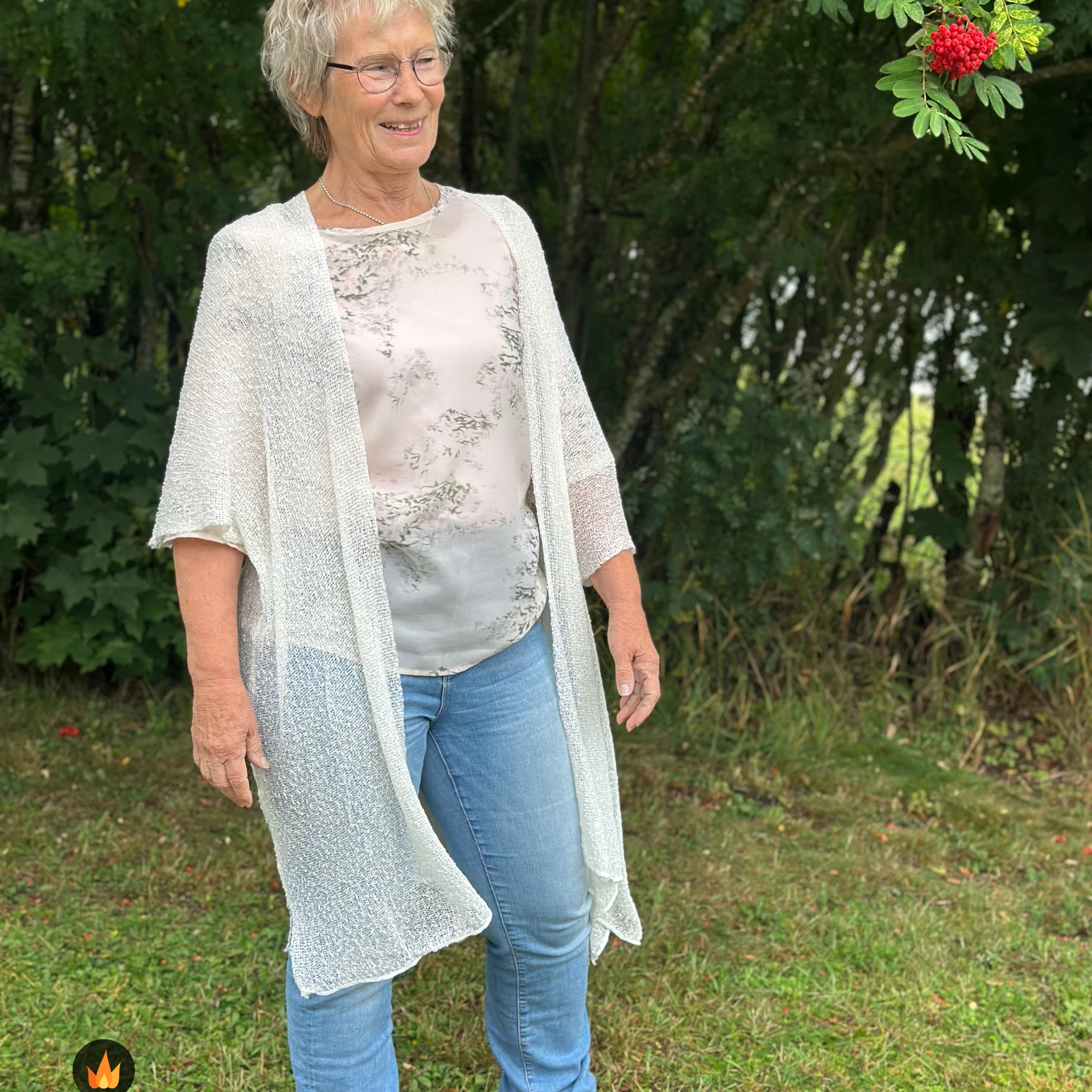 Kvinna bär Biasa Kofta i färg 031 Offwhite, en varm färg i mjuk offwhite, lång finstickad oversized kofta buren öppen, fotad utomhus i grönska. Ulla Jacobsson Kläder.