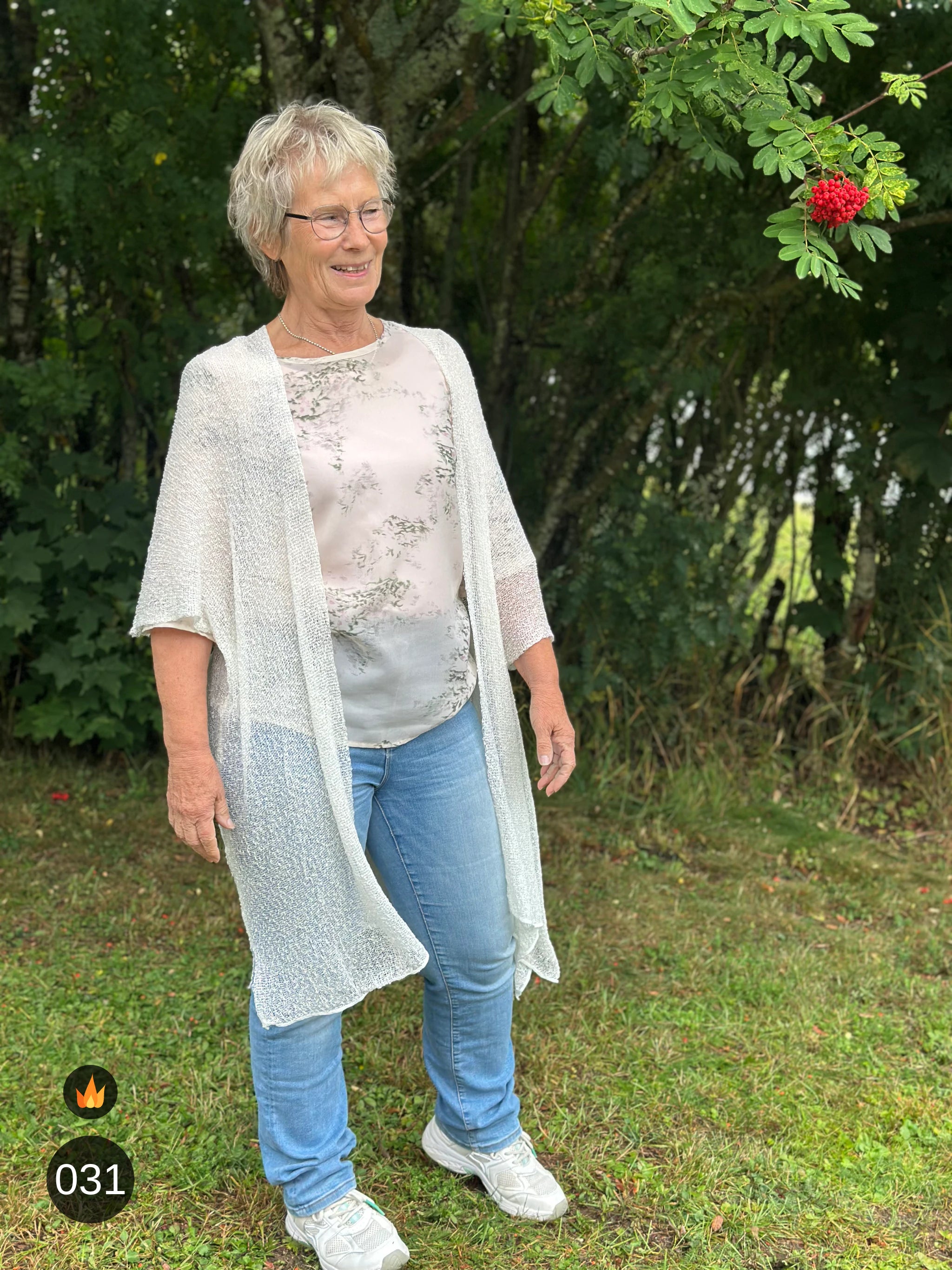 Kvinna bär Biasa Kofta i färg 031 Offwhite, en varm färg i mjuk offwhite, lång finstickad oversized kofta buren öppen, fotad utomhus i grönska. Ulla Jacobsson Kläder.
