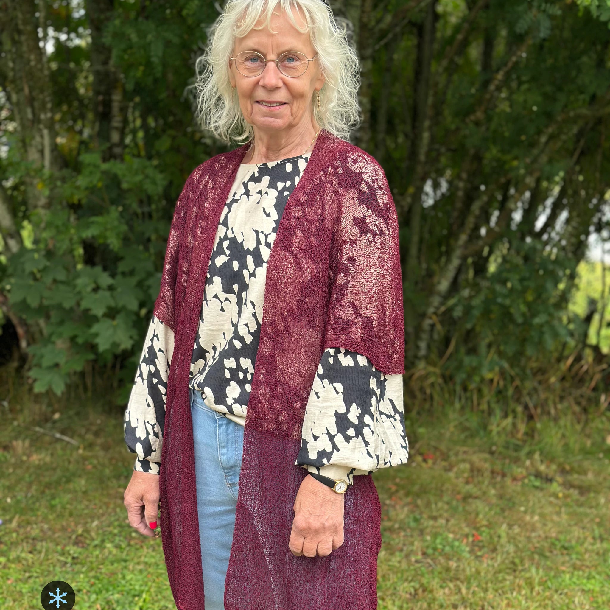 Kvinna bär Biasa Kofta i färg 047 Bourdeaux, en kall färg i djup bourdeauxröd, lång finstickad oversized kofta buren öppen, fotad utomhus i grönska. Ulla Jacobsson Kläder.