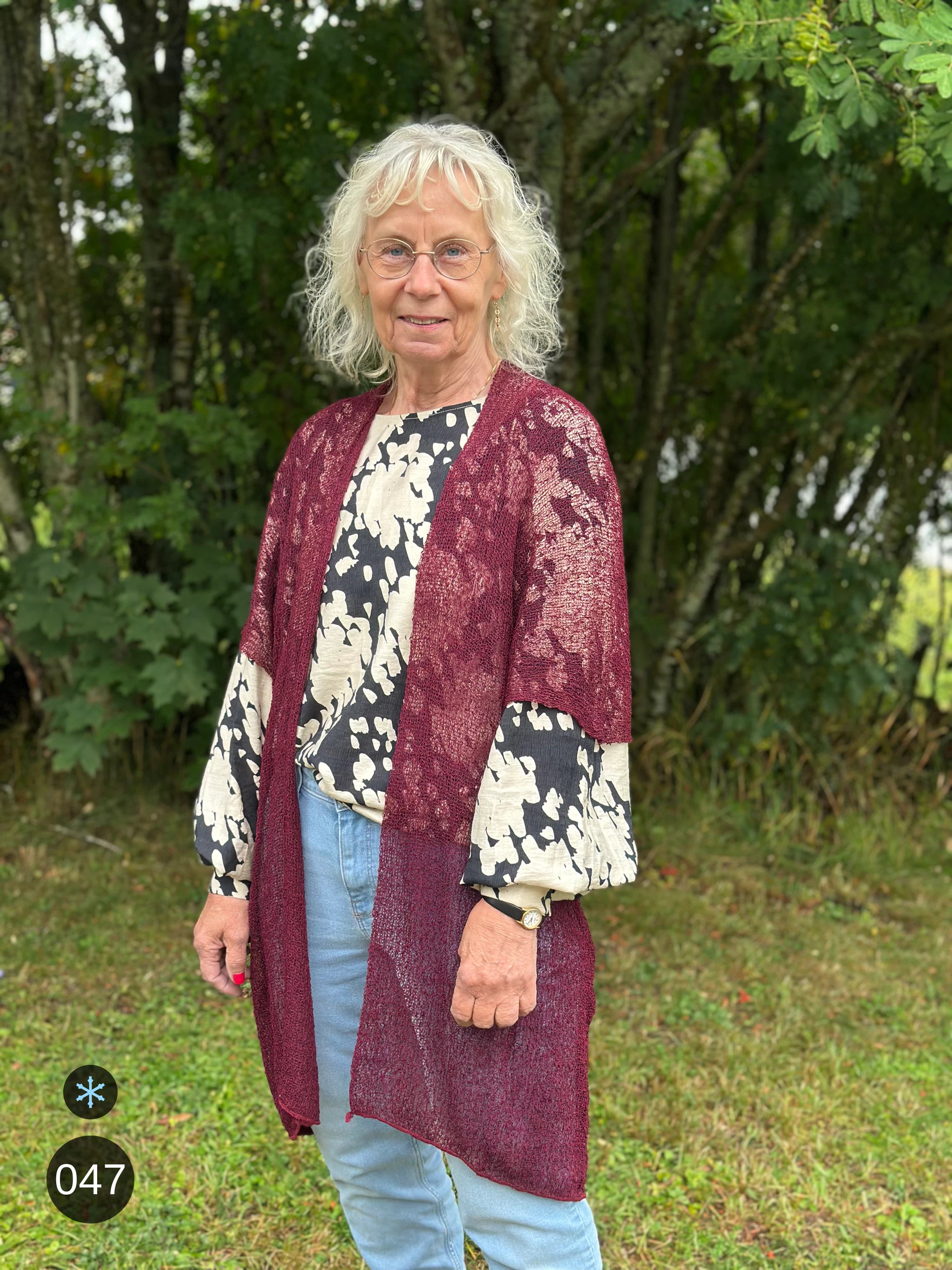 Kvinna bär Biasa Kofta i färg 047 Bourdeaux, en kall färg i djup bourdeauxröd, lång finstickad oversized kofta buren öppen, fotad utomhus i grönska. Ulla Jacobsson Kläder.