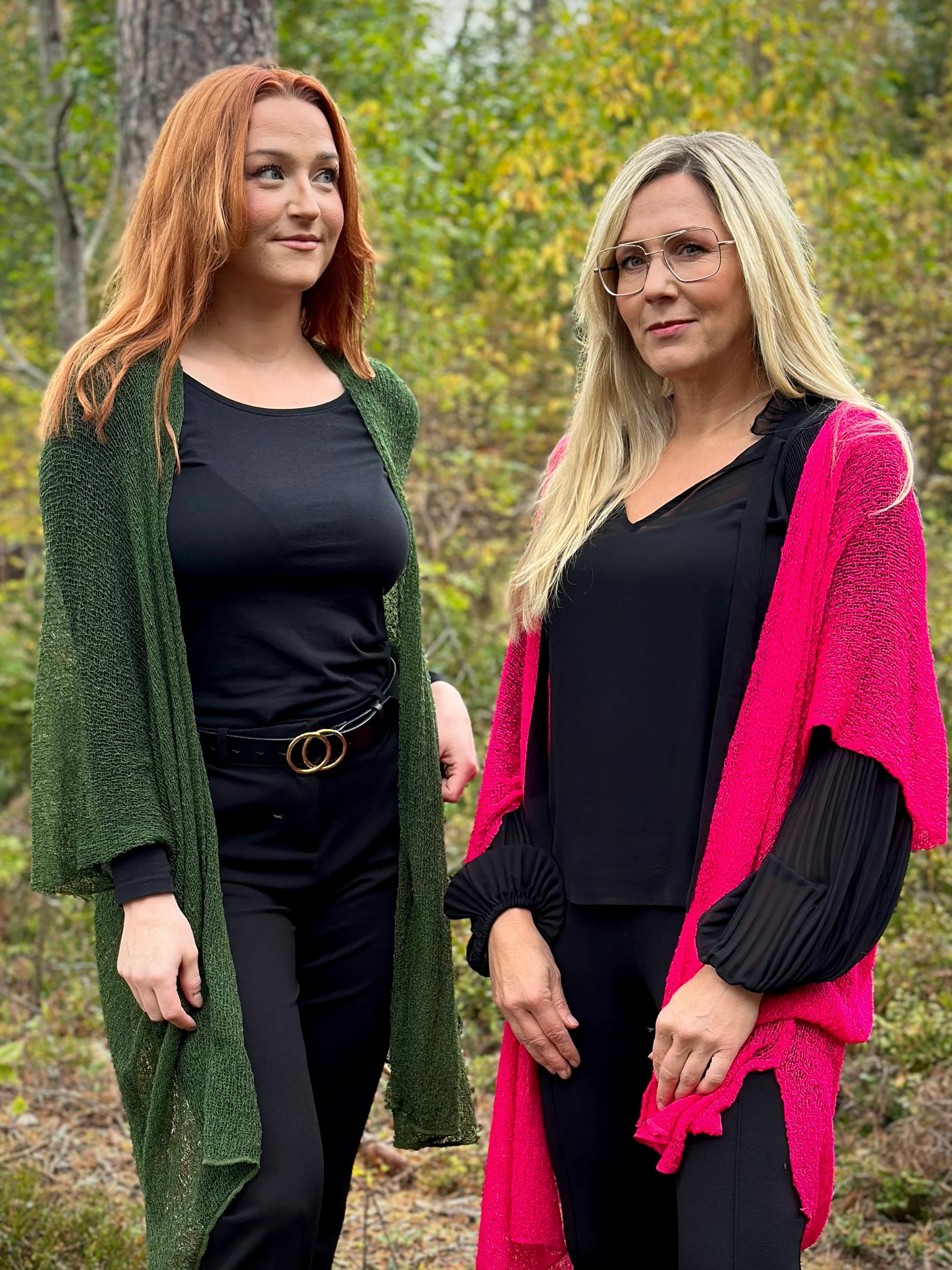 Två kvinnor bär Biasa Kofta, den ena i 061 Mörkgrön och den andra i 070 Neonrosa. Långa finstickade oversized koftor burna öppna, fotade i höstskog. Ulla Jacobsson Kläder.