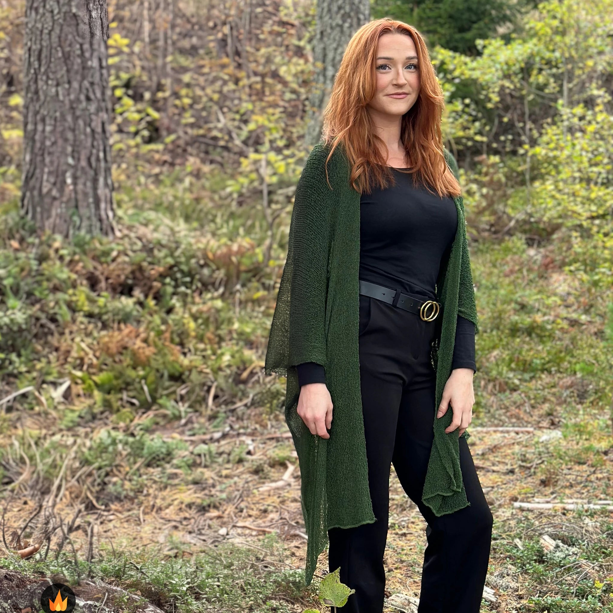 Kvinna bär Biasa Kofta i färg 061 Mörkgrön, en varm färg i djup mörkgrön, lång finstickad oversized kofta buren öppen, fotad i höstskog bland tallar. Ulla Jacobsson Kläder.