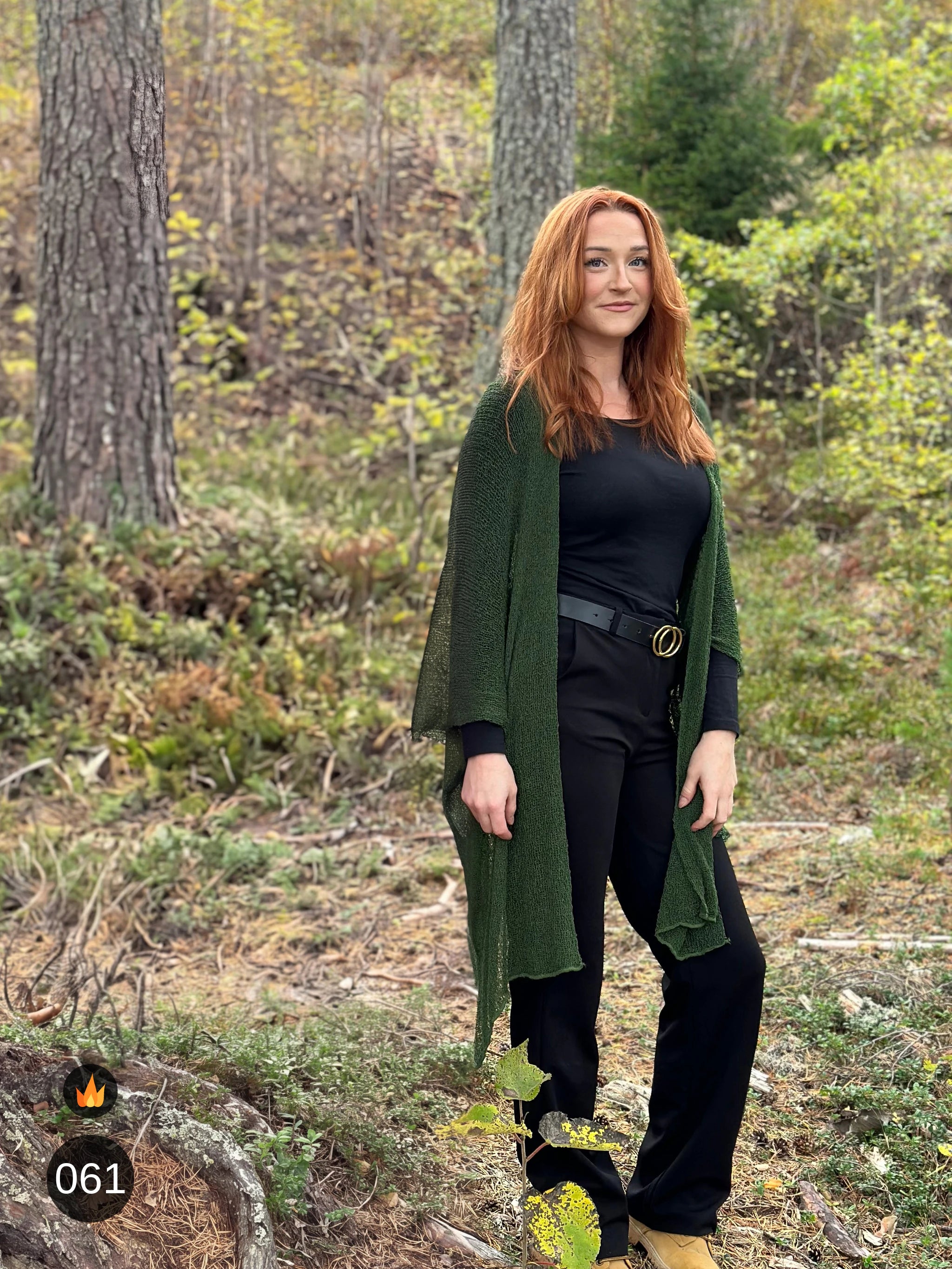 Kvinna bär Biasa Kofta i färg 061 Mörkgrön, en varm färg i djup mörkgrön, lång finstickad oversized kofta buren öppen, fotad i höstskog bland tallar. Ulla Jacobsson Kläder.