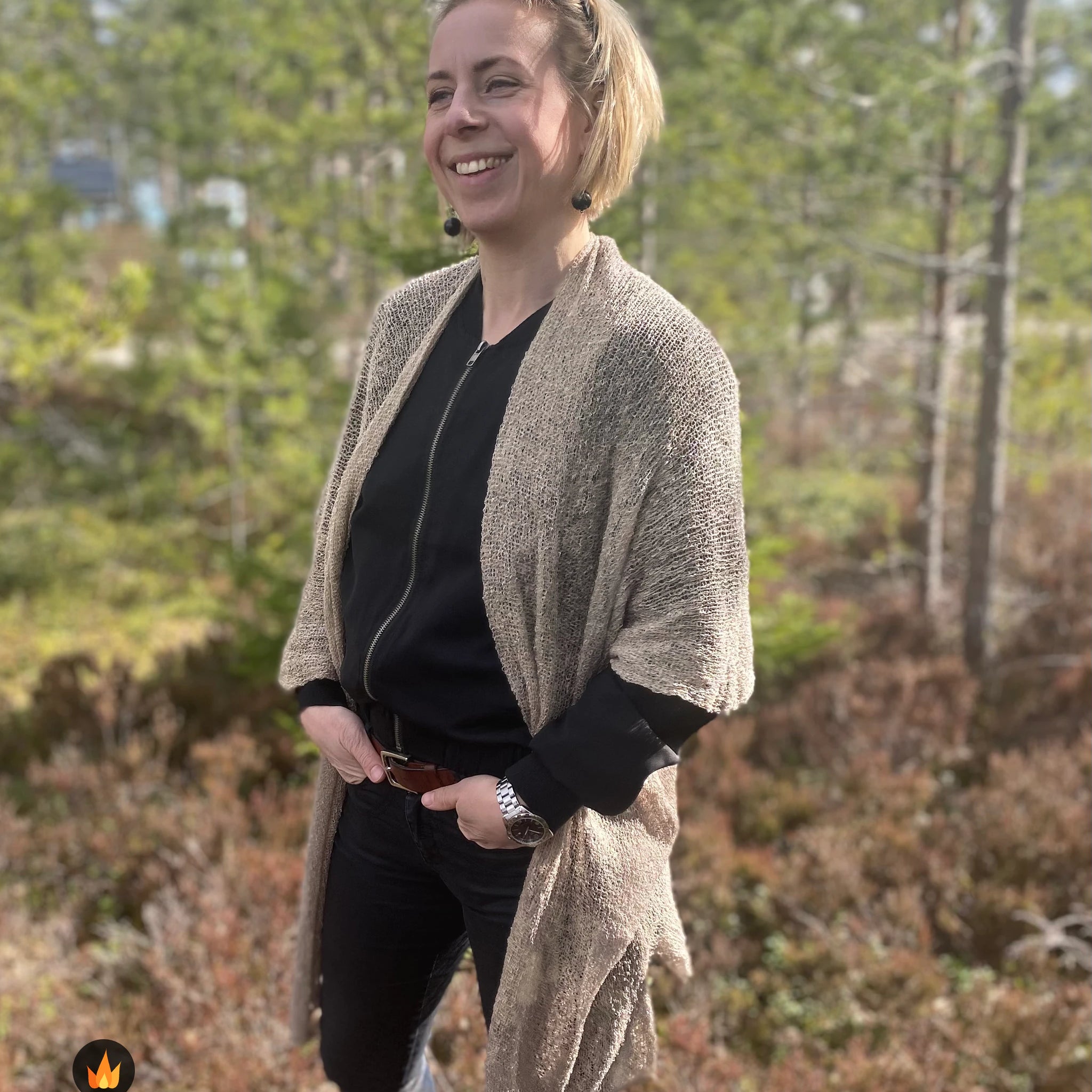 Nainen musta huivi päässään vaaleaa seinää vasten yllään vaalean beige Biasa-neuletakki; pehmeä, ohutneulottu takki asettuu hartioille vaalean harmaanbeigenä. Ulla Jacobsson Kläder