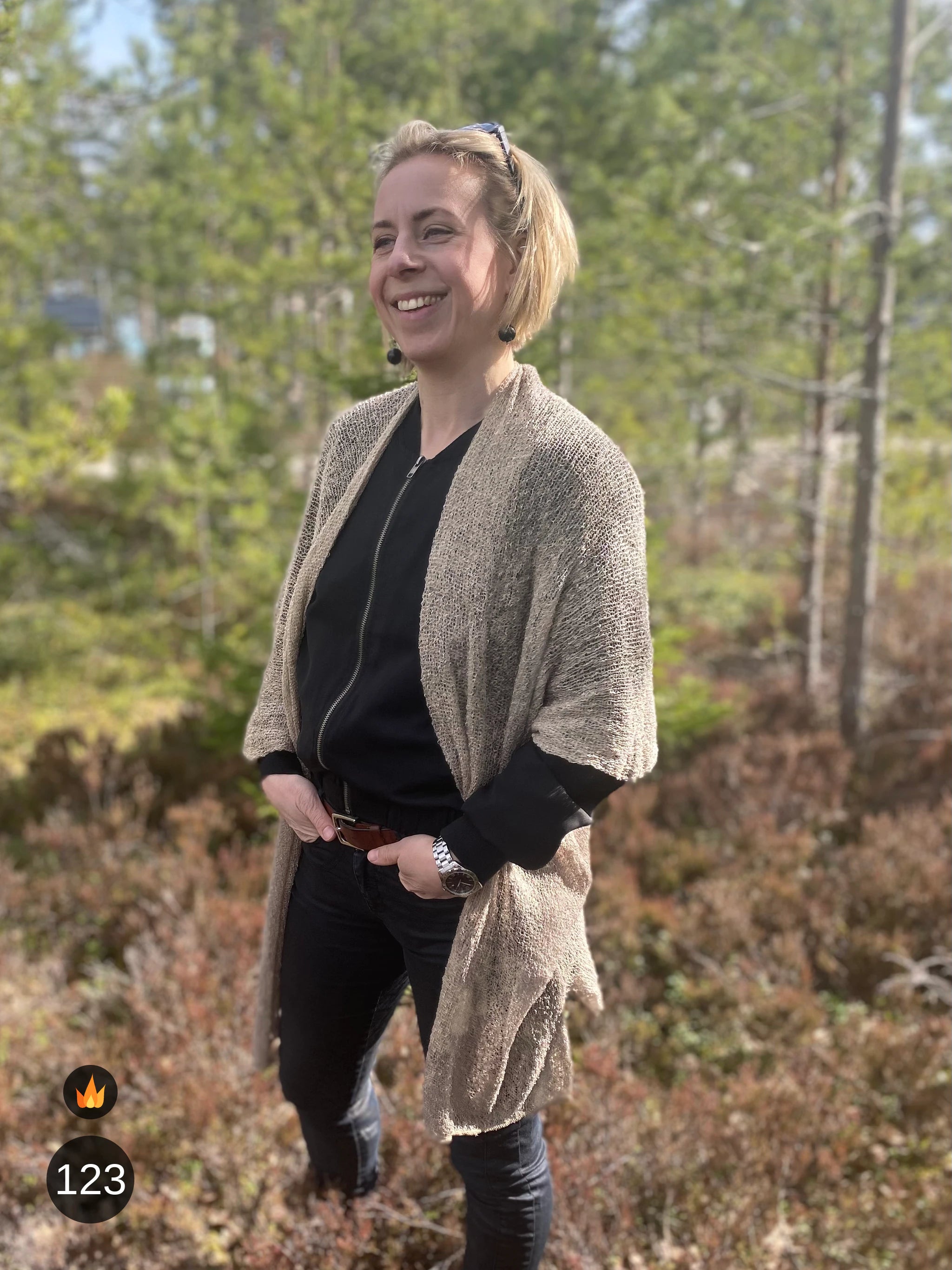 Nainen musta huivi päässään vaaleaa seinää vasten yllään vaalean beige Biasa-neuletakki; pehmeä, ohutneulottu takki asettuu hartioille vaalean harmaanbeigenä. Ulla Jacobsson Kläder