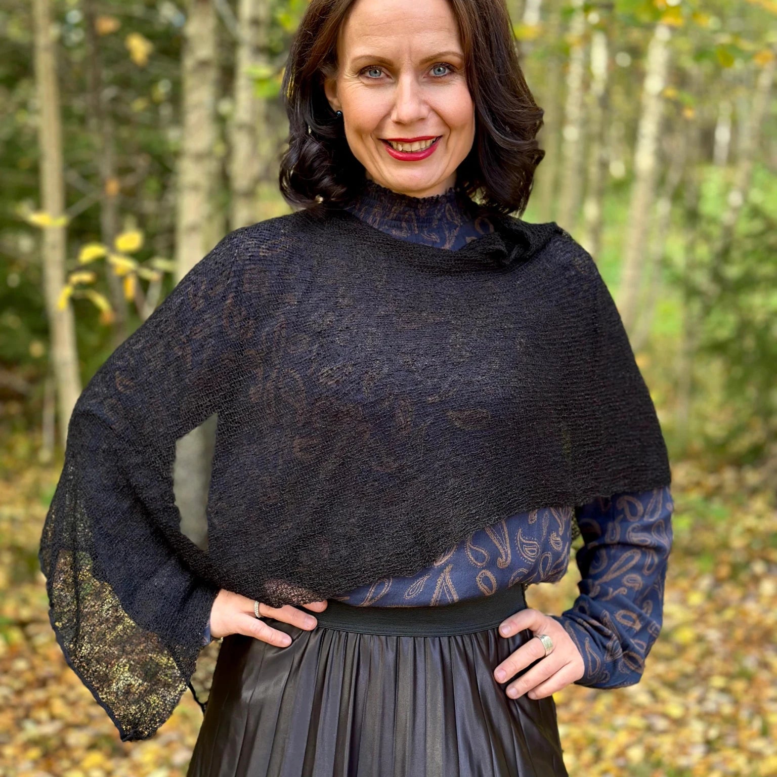 Kvinna bär Ella Cape i färg 001 Svart, en neutral djupsvart med matt yta, finstickad cape som draperas mjukt över axlarna, fotad i en höstskog med gula björklöv. Ulla Jacobsson Kläder.