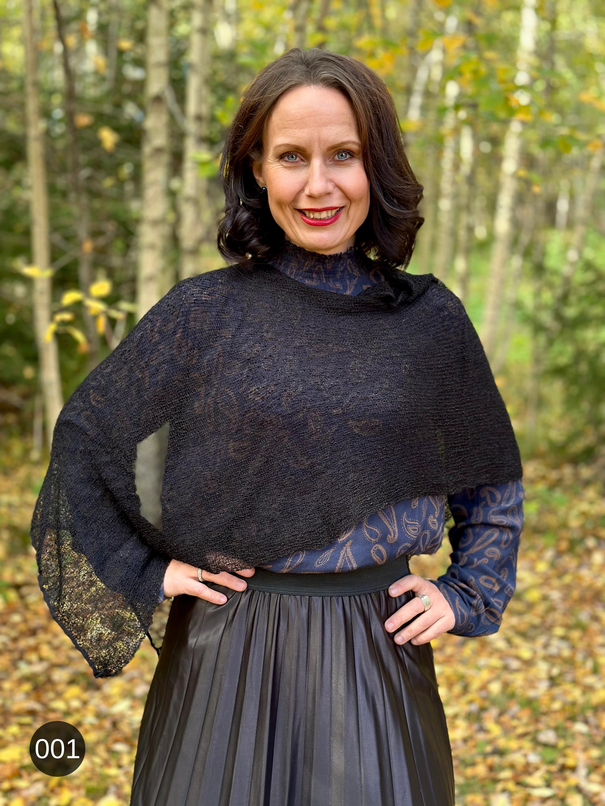Kvinna bär Ella Cape i färg 001 Svart, en neutral djupsvart med matt yta, finstickad cape som draperas mjukt över axlarna, fotad i en höstskog med gula björklöv. Ulla Jacobsson Kläder.