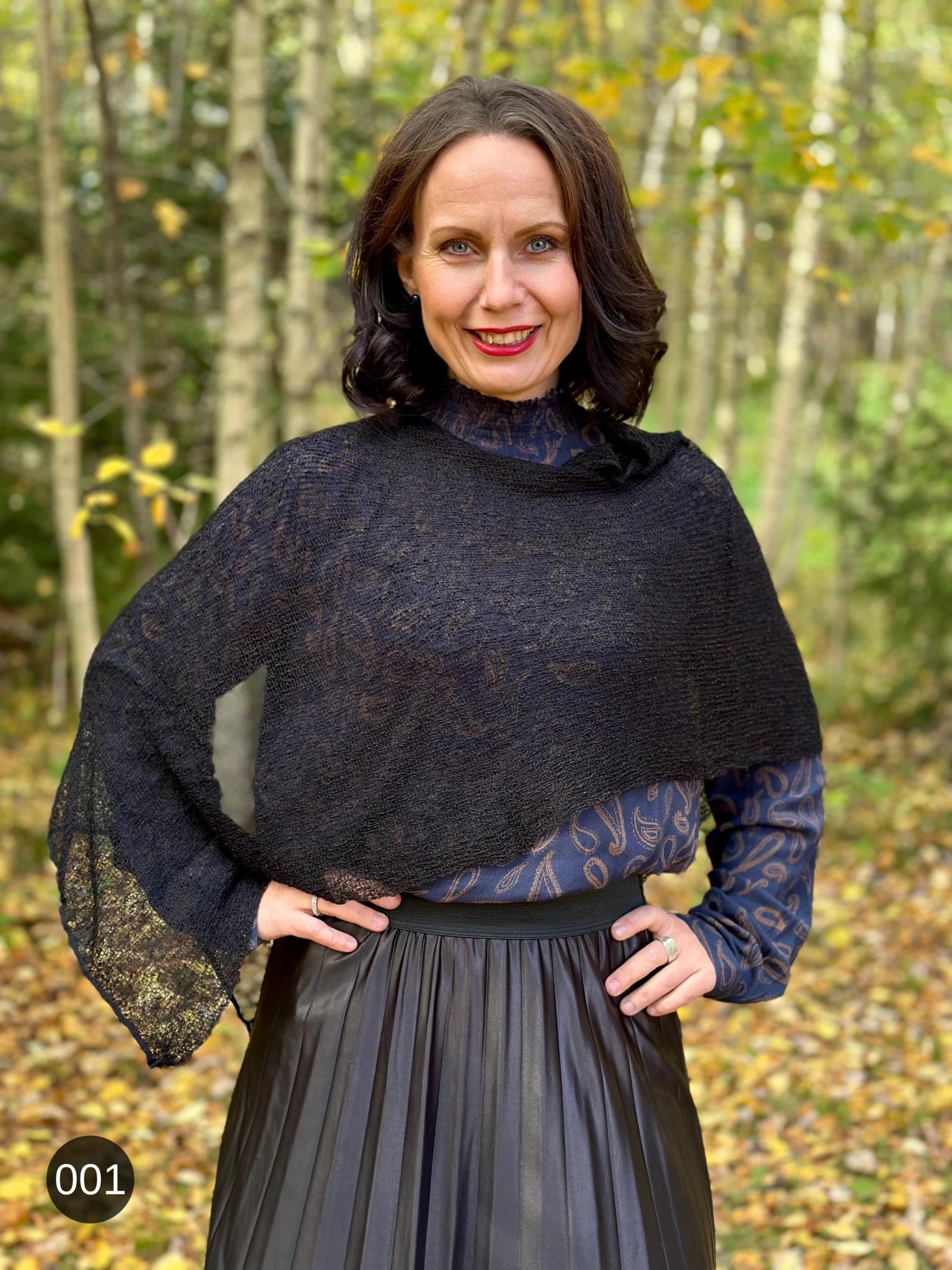 Kvinna bär Ella Cape i färg 001 Svart, en neutral djupsvart med matt yta, finstickad cape som draperas mjukt över axlarna, fotad i en höstskog med gula björklöv. Ulla Jacobsson Kläder.
