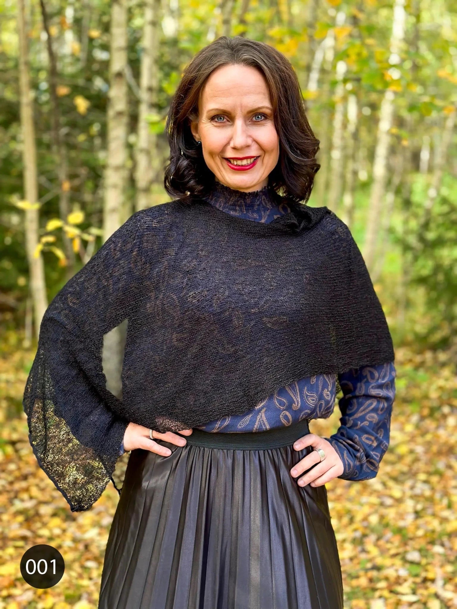 I en höstskog med gula löv står en kvinna med handen i sidan i en svart Ella cape, den glesa stickningen ramar in axlarna och kanten faller ned över en mörk plisserad kjol. Ulla Jacobsson Kläder