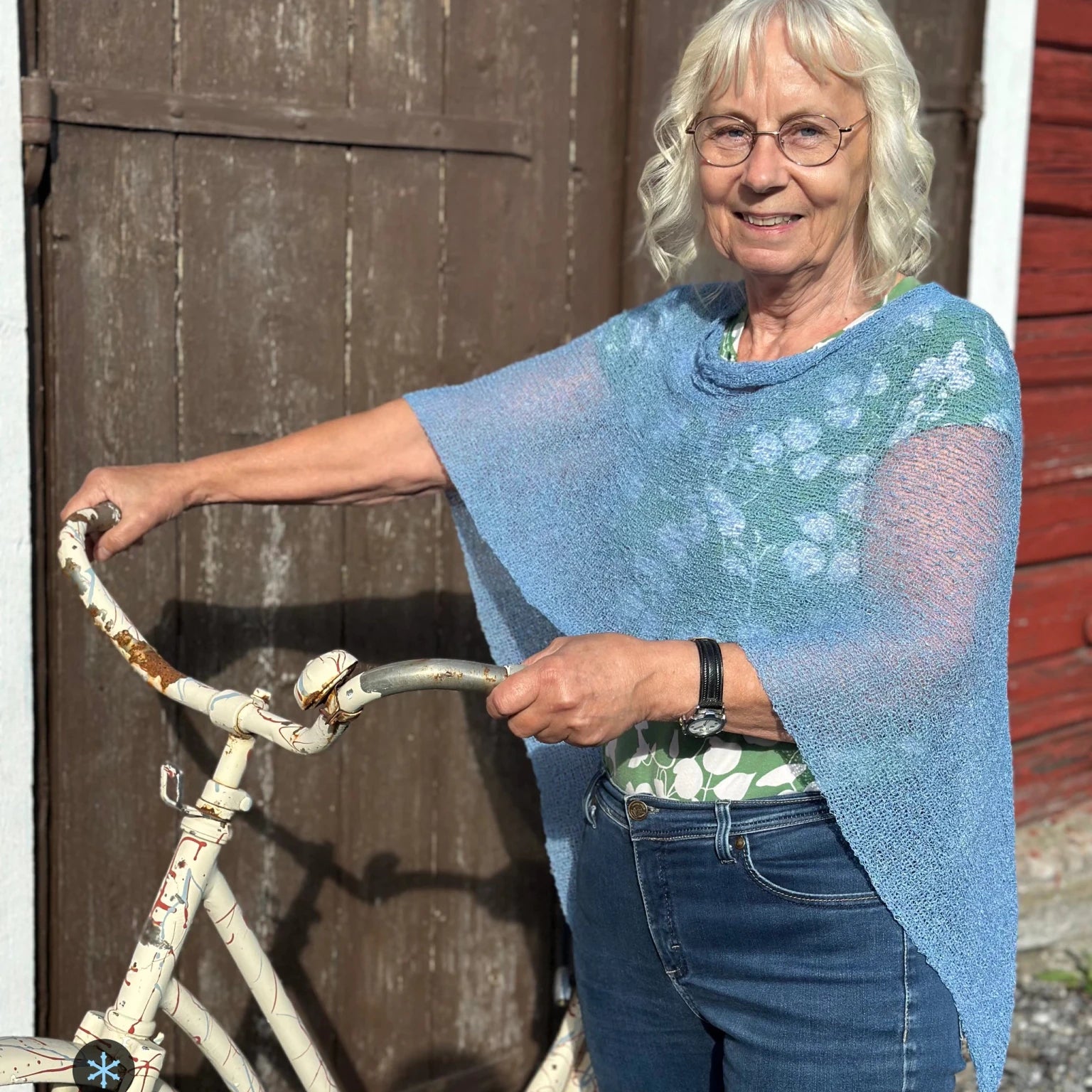 Kvinna bär Ella Cape i färg 003 Isblå, en kall färg i ljus, sval isblå med silvrig dager, finstickad cape som draperas mjukt över axlarna, fotad vid en faluröd ladvägg med en vit cykel. Ulla Jacobsson Kläder.
