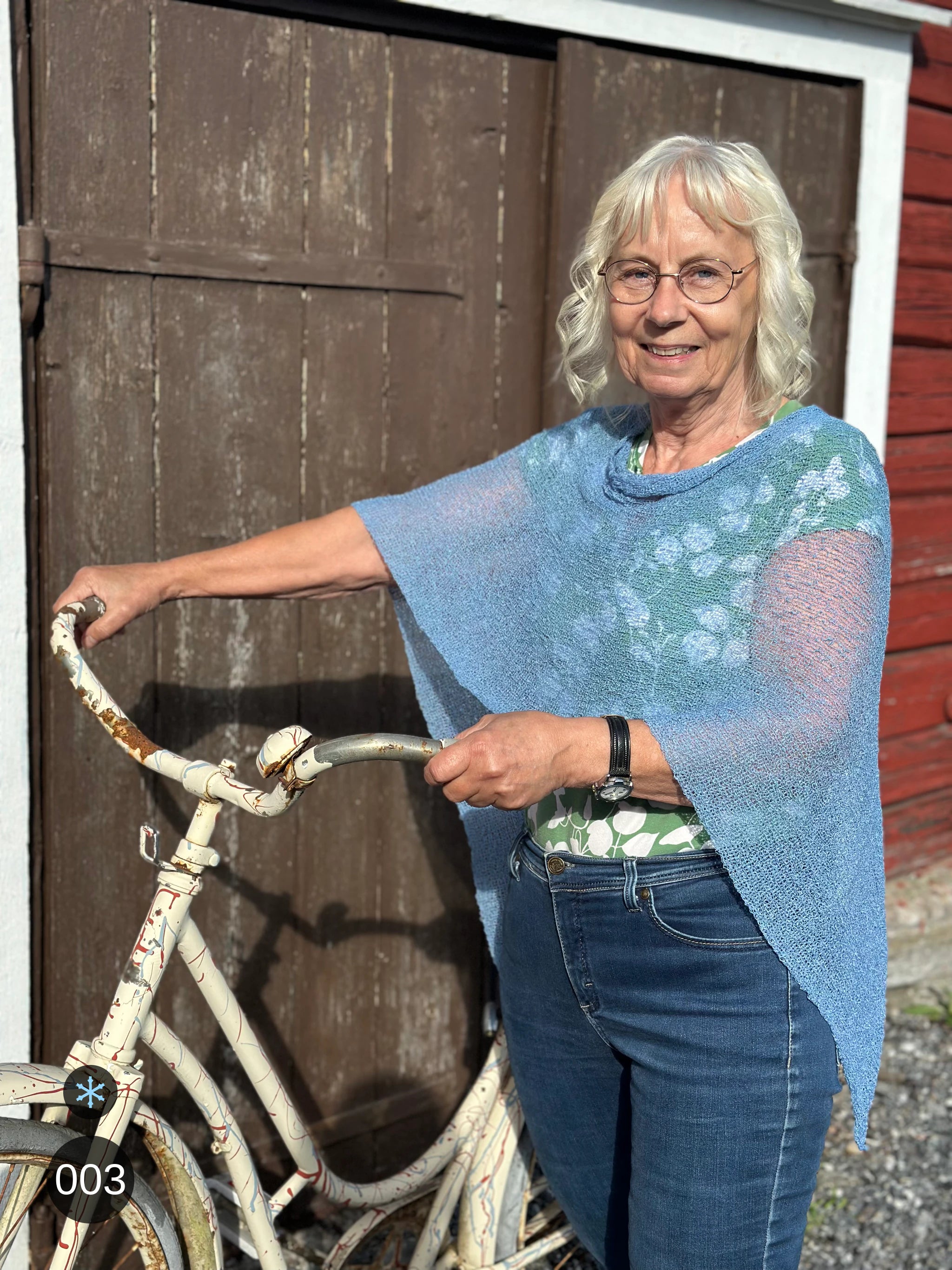 Kvinna bär Ella Cape i färg 003 Isblå, en kall färg i ljus, sval isblå med silvrig dager, finstickad cape som draperas mjukt över axlarna, fotad vid en faluröd ladvägg med en vit cykel. Ulla Jacobsson Kläder.