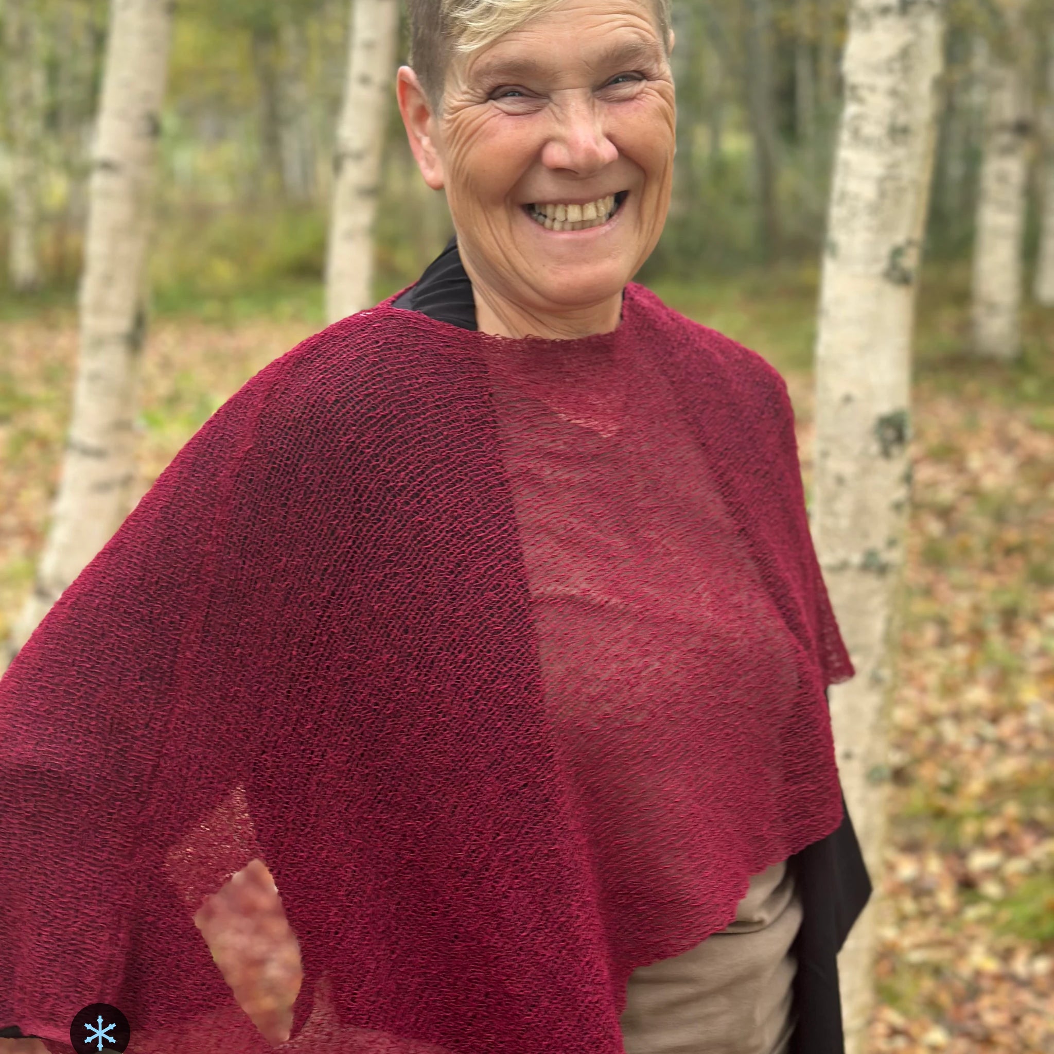 Kvinna bär Ella Cape i färg 005 Vinröd, en kall färg i djup vinröd med blå underton, finstickad cape som draperas mjukt över axlarna, fotad bland björkar en höstdag. Ulla Jacobsson Kläder.