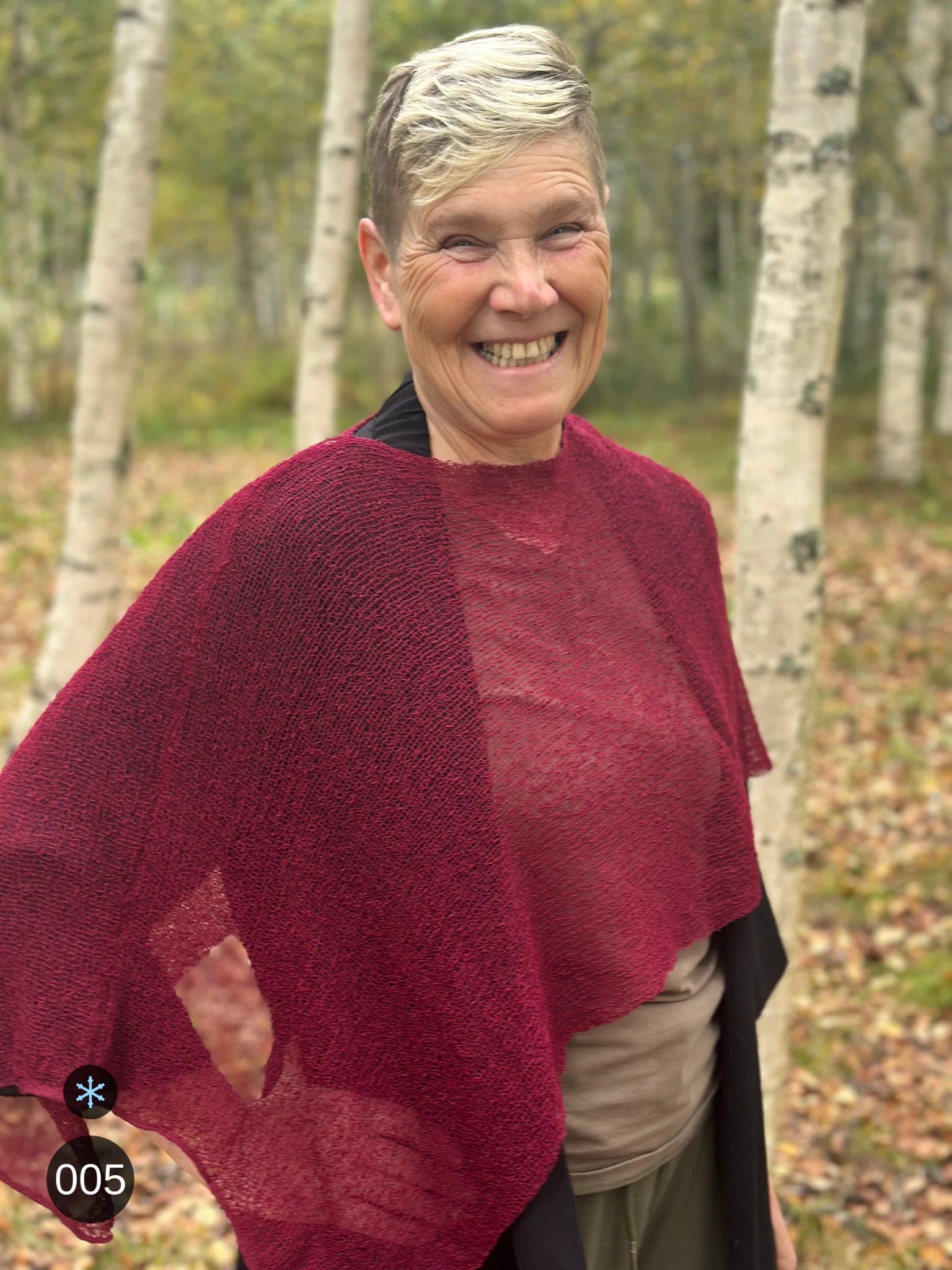 Kvinna bär Ella Cape i färg 005 Vinröd, en kall färg i djup vinröd med blå underton, finstickad cape som draperas mjukt över axlarna, fotad bland björkar en höstdag. Ulla Jacobsson Kläder.