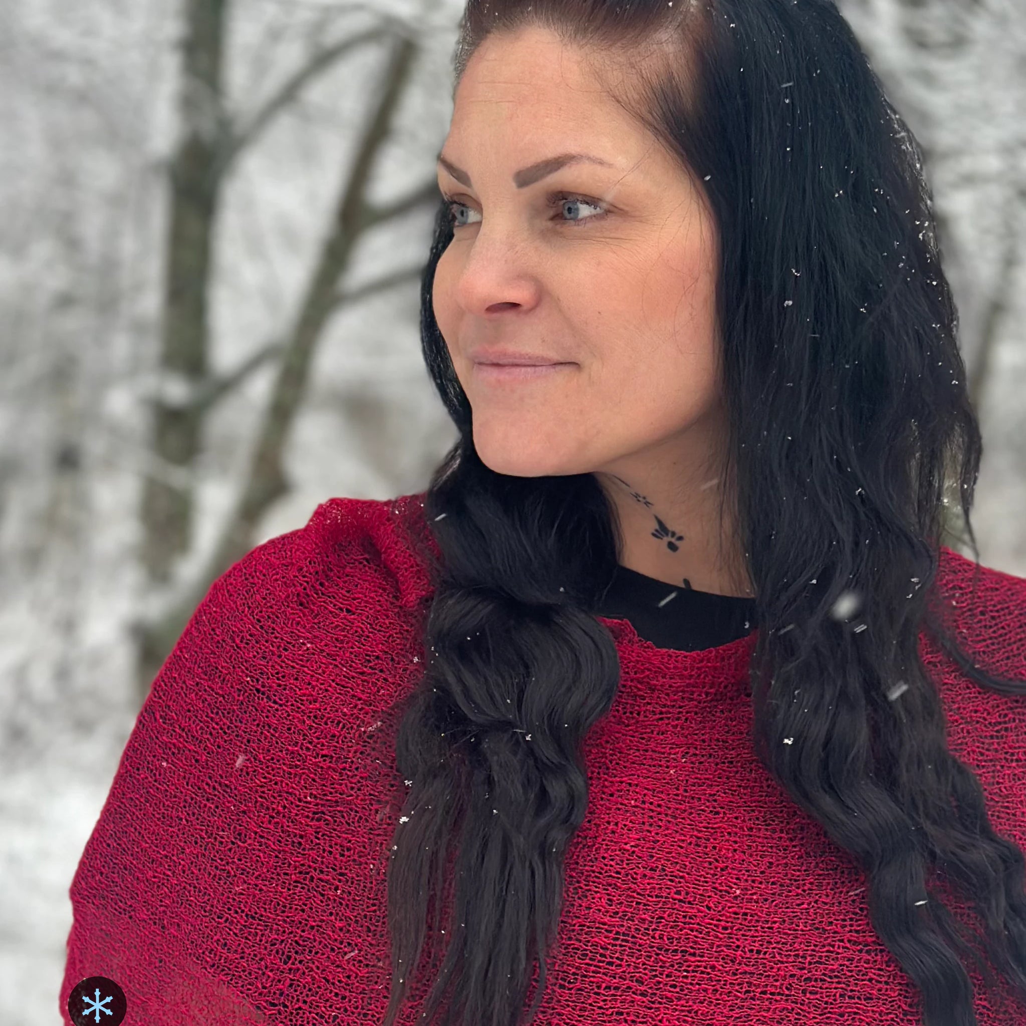 Kvinna bär Ella Cape i färg 006 Chili, en kall färg i djup chiliröd med blå underton, finstickad cape som draperas mjukt över axlarna, fotad i lätt snöfall bland björkar. Ulla Jacobsson Kläder.