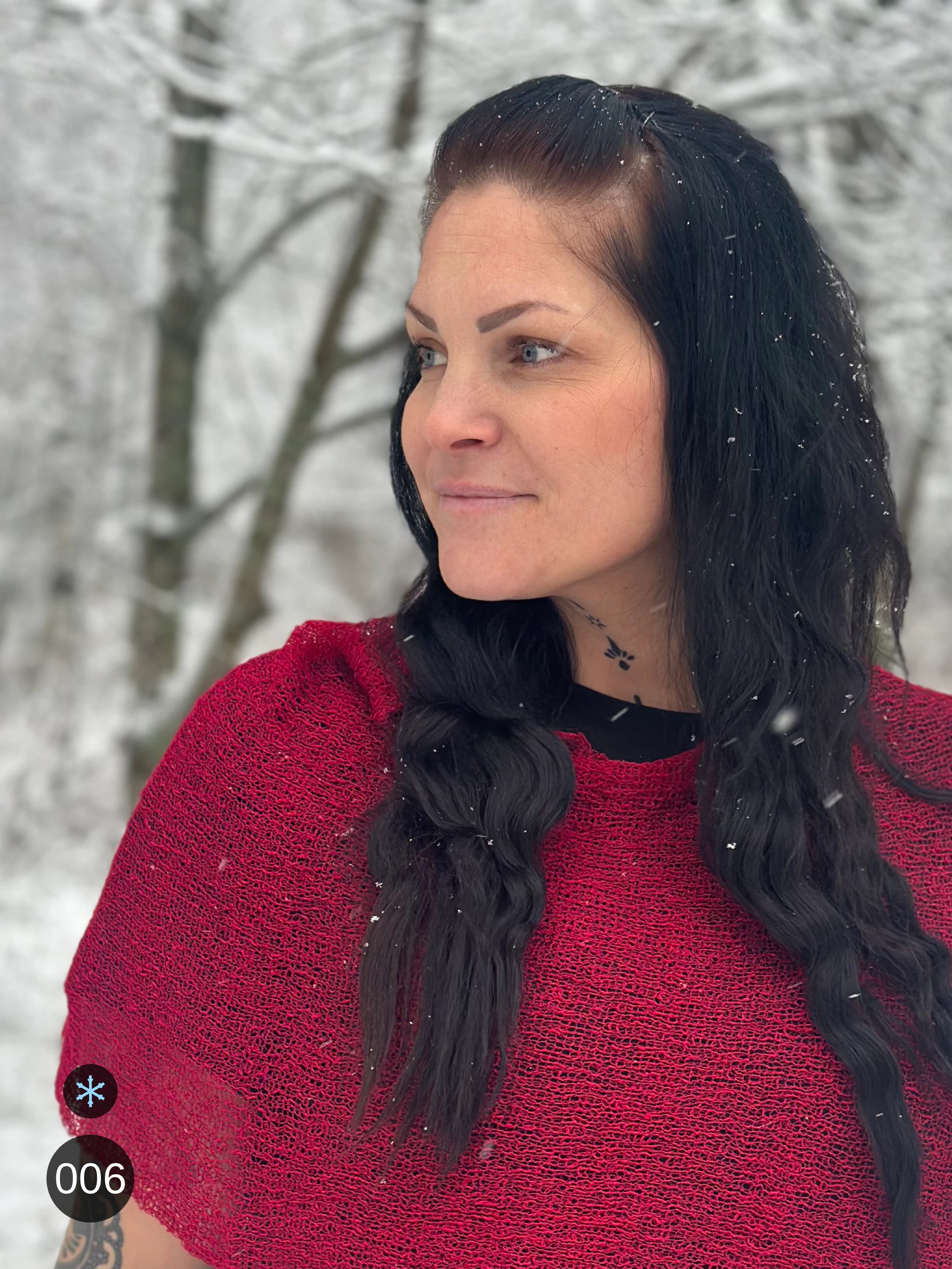 Kvinna bär Ella Cape i färg 006 Chili, en kall färg i djup chiliröd med blå underton, finstickad cape som draperas mjukt över axlarna, fotad i lätt snöfall bland björkar. Ulla Jacobsson Kläder.