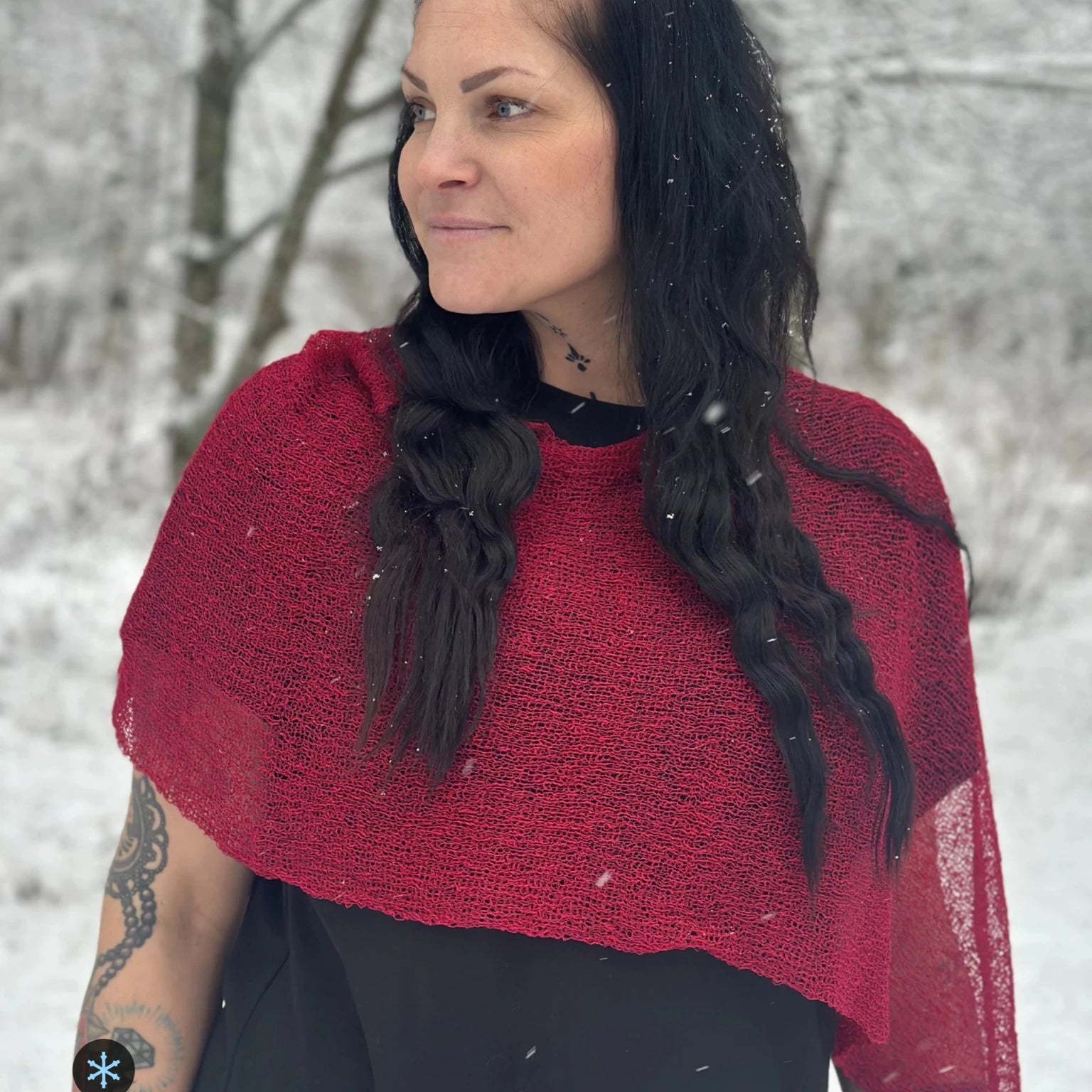 Kvinna bär Ella Cape i färg 006 Chili, en kall färg, finstickad cape som draperas mjukt över axlarna, fotad i lätt snöfall bland björkar. Ulla Jacobsson Kläder.