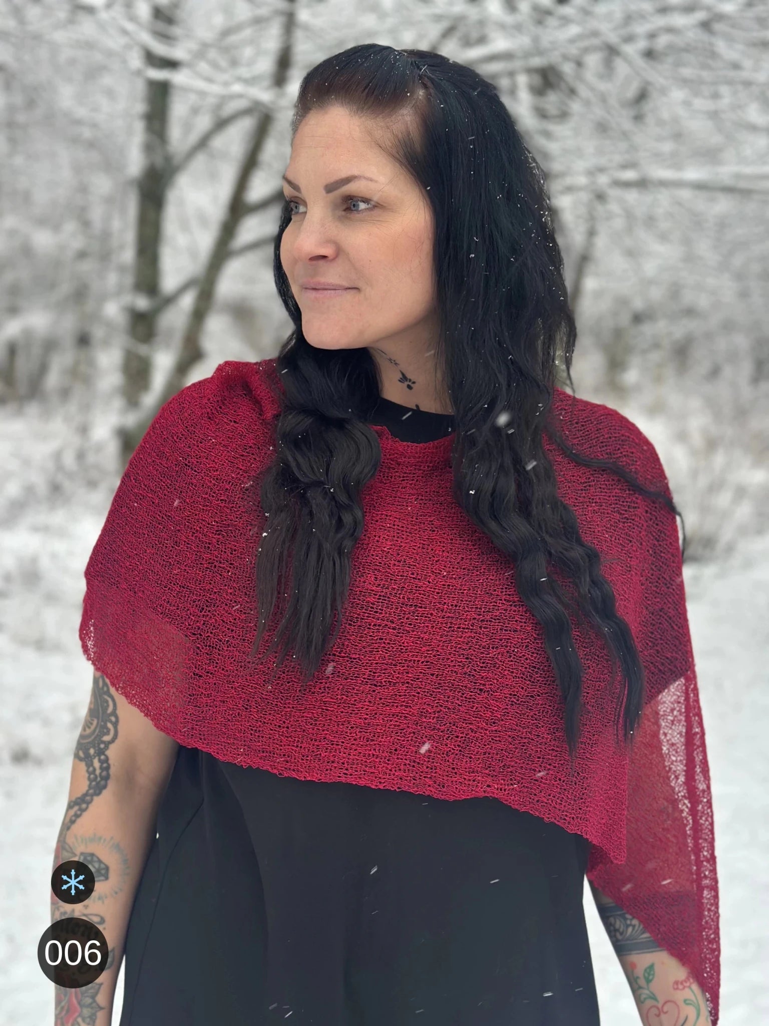 Kvinna bär Ella Cape i färg 006 Chili, en kall färg, finstickad cape som draperas mjukt över axlarna, fotad i lätt snöfall bland björkar. Ulla Jacobsson Kläder.