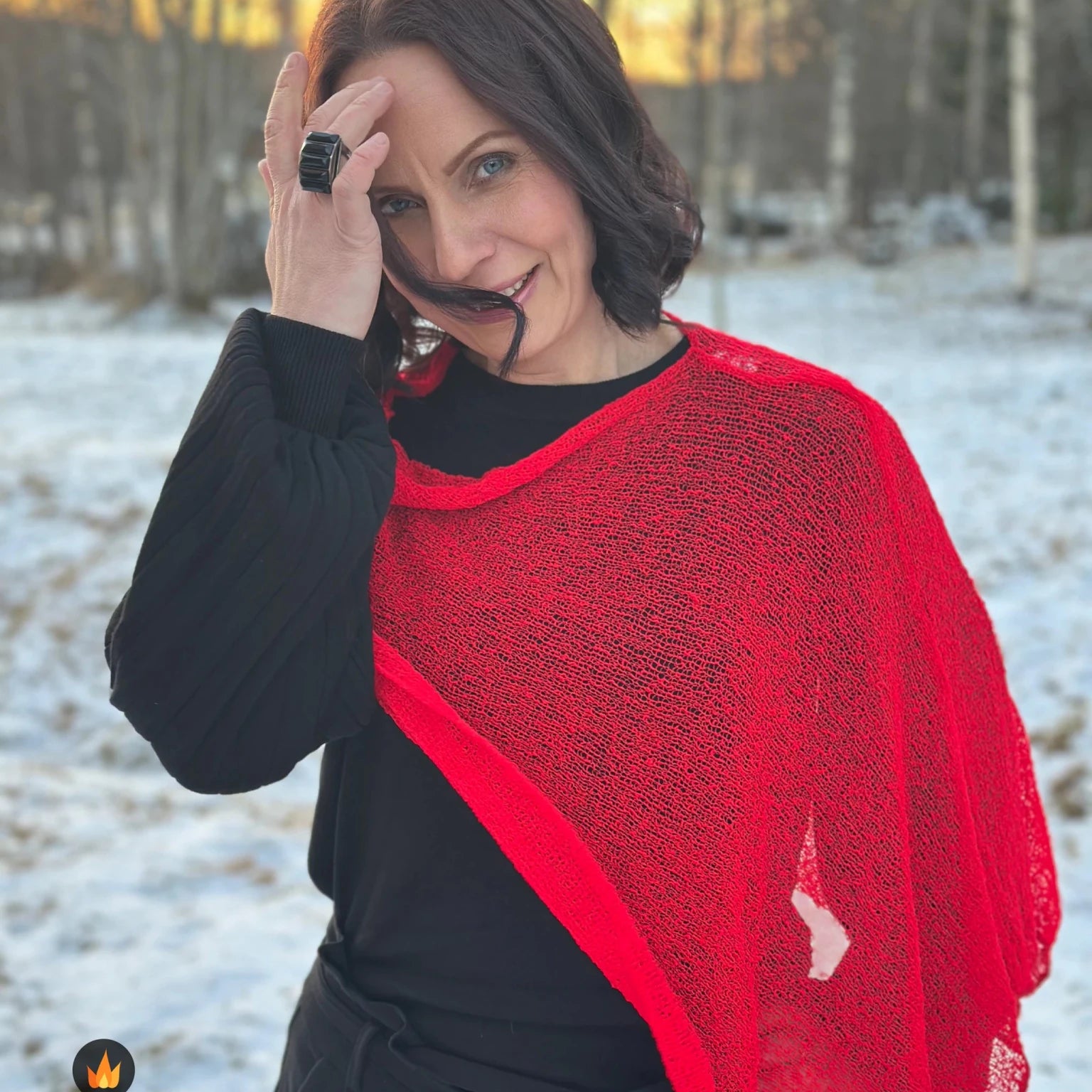 Kvinna bär Ella Cape i färg 007 Klarröd, en varm färg i klar signalröd med lätt orange glöd, finstickad cape som draperas mjukt över axlarna, fotad i snöigt vinterljus. Ulla Jacobsson Kläder.