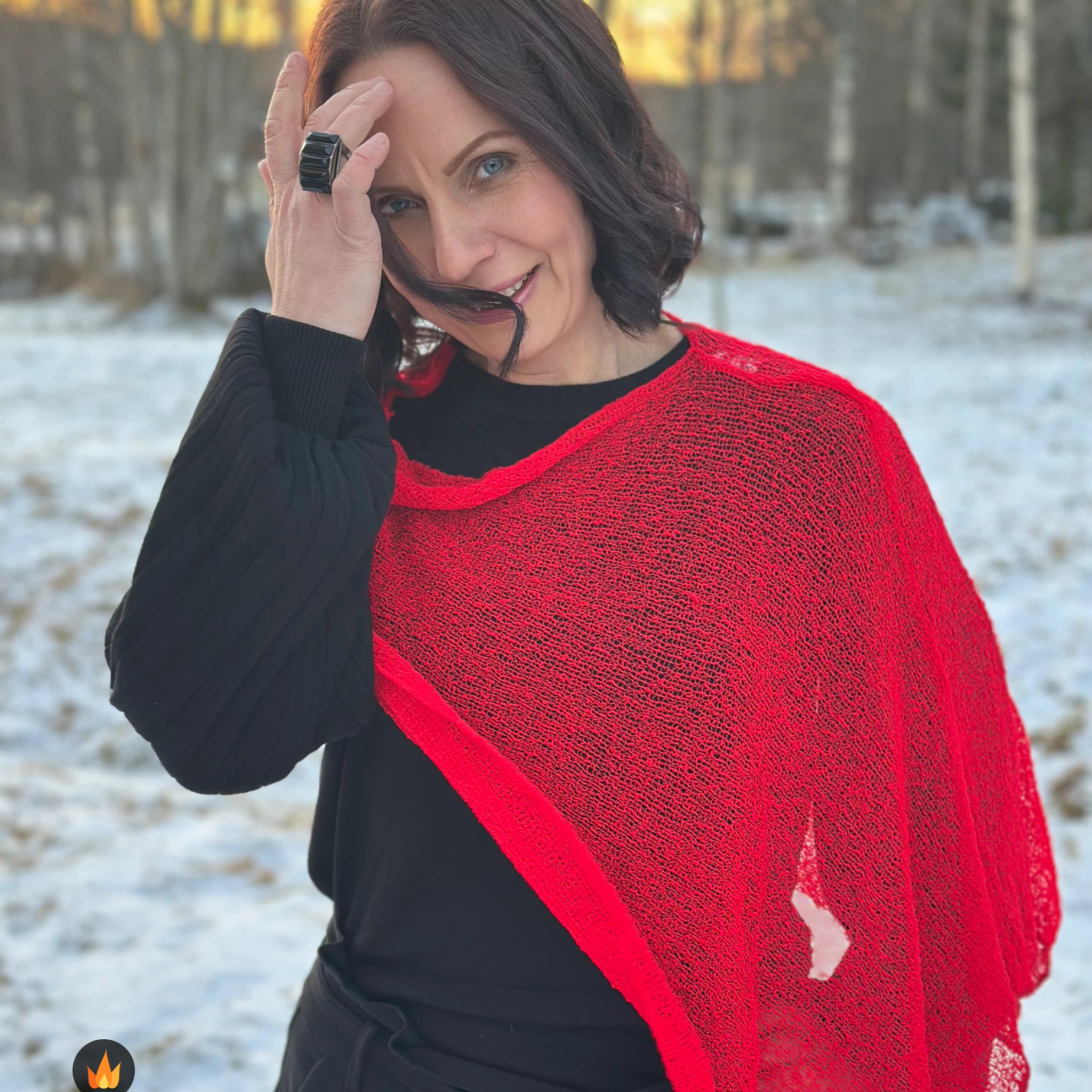 Kvinna bär Ella Cape i färg 007 Klarröd, en varm färg i klar signalröd med lätt orange glöd, finstickad cape som draperas mjukt över axlarna, fotad i snöigt vinterljus. Ulla Jacobsson Kläder.