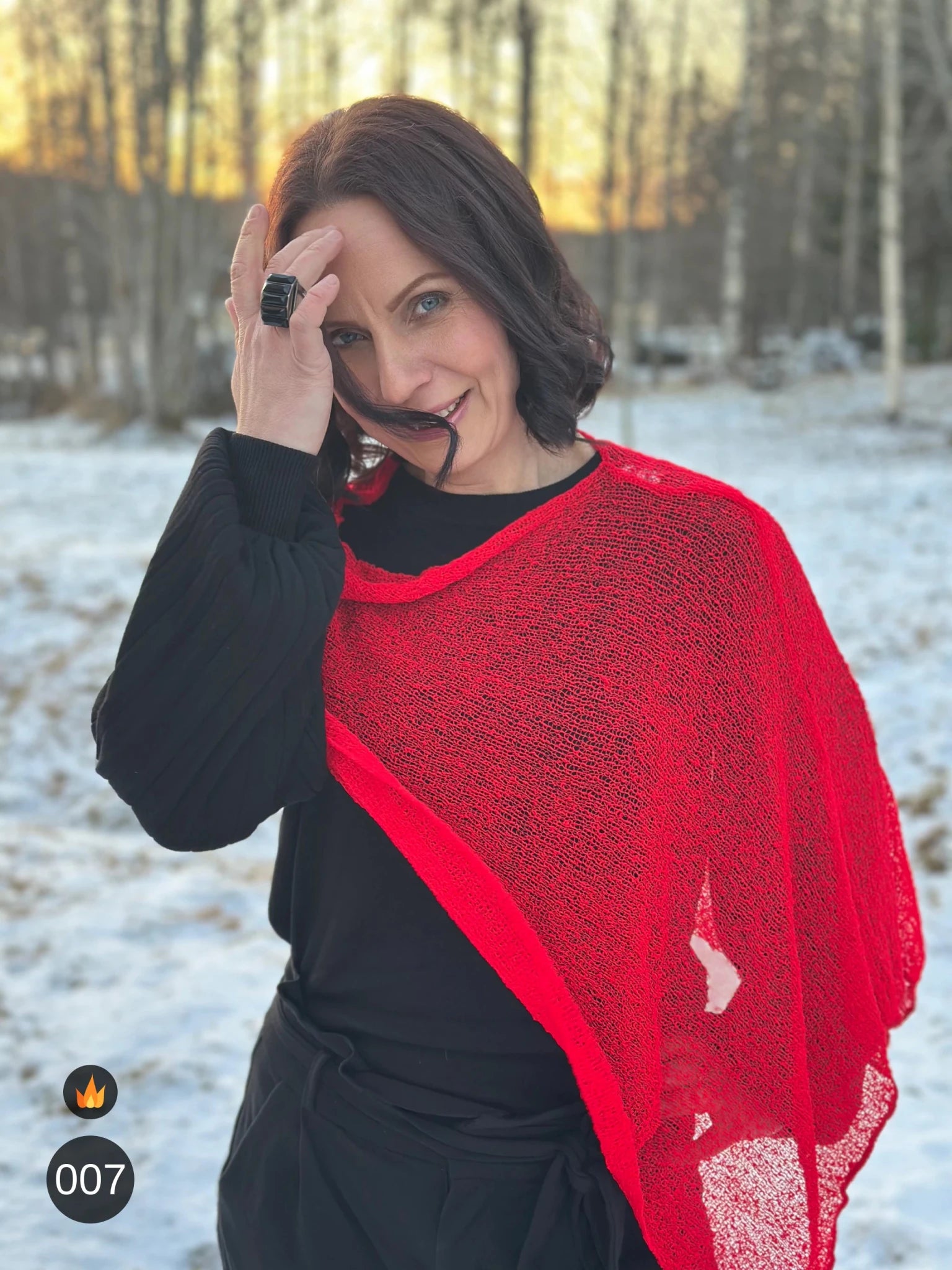 Kvinna bär Ella Cape i färg 007 Klarröd, en varm färg i klar signalröd med lätt orange glöd, finstickad cape som draperas mjukt över axlarna, fotad i snöigt vinterljus. Ulla Jacobsson Kläder.