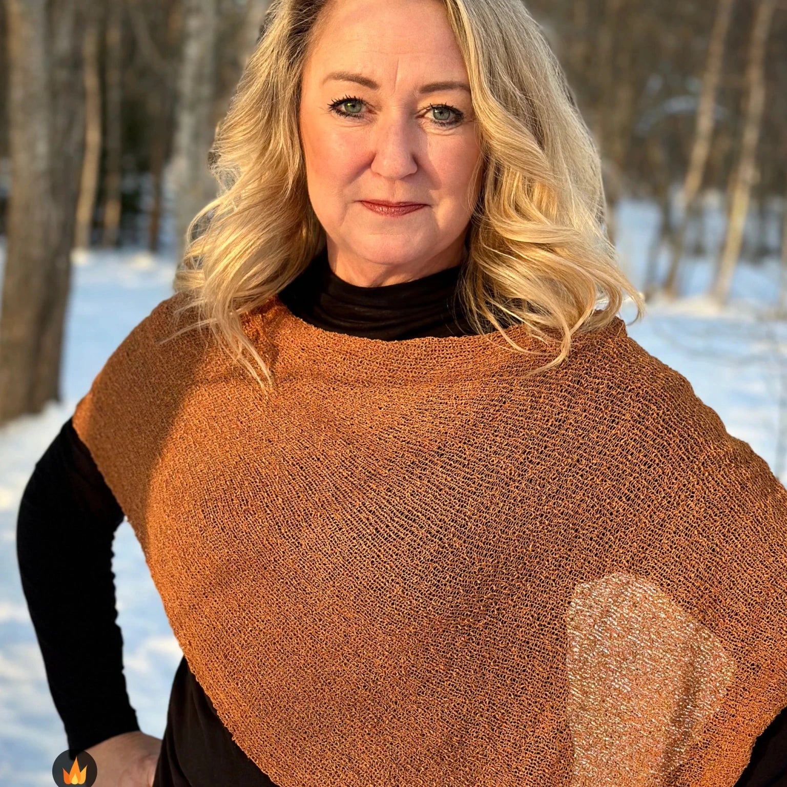 Kvinna bär Ella Cape i färg 009 Mocka, en varm färg i mockabrun med kopparglöd, finstickad cape som draperas mjukt över axlarna, fotad i vintersol vid snötäckta björkar. Ulla Jacobsson Kläder.