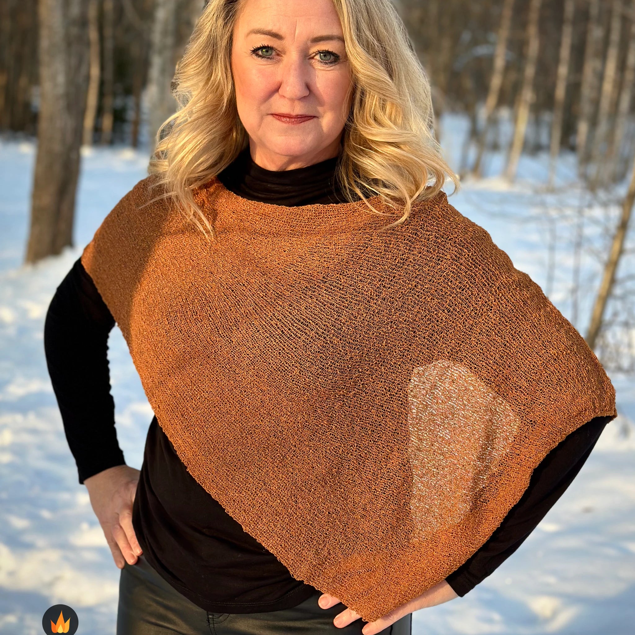 Kvinna bär Ella Cape i färg 009 Mocka, en varm färg i mockabrun med kopparglöd, finstickad cape som draperas mjukt över axlarna, fotad i vintersol vid snötäckta björkar. Ulla Jacobsson Kläder.