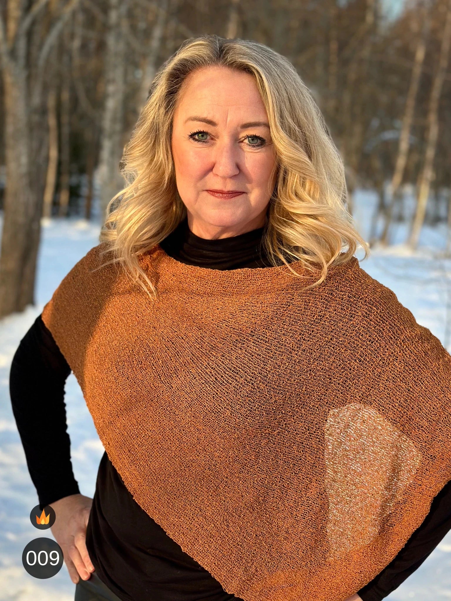 Kvinna bär Ella Cape i färg 009 Mocka, en varm färg i mockabrun med kopparglöd, finstickad cape som draperas mjukt över axlarna, fotad i vintersol vid snötäckta björkar. Ulla Jacobsson Kläder.