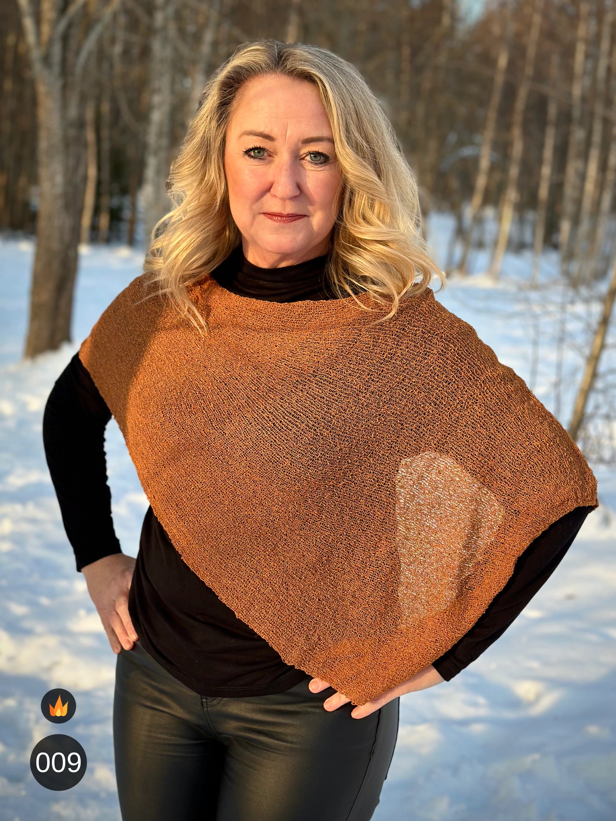 Kvinna bär Ella Cape i färg 009 Mocka, en varm färg i mockabrun med kopparglöd, finstickad cape som draperas mjukt över axlarna, fotad i vintersol vid snötäckta björkar. Ulla Jacobsson Kläder.