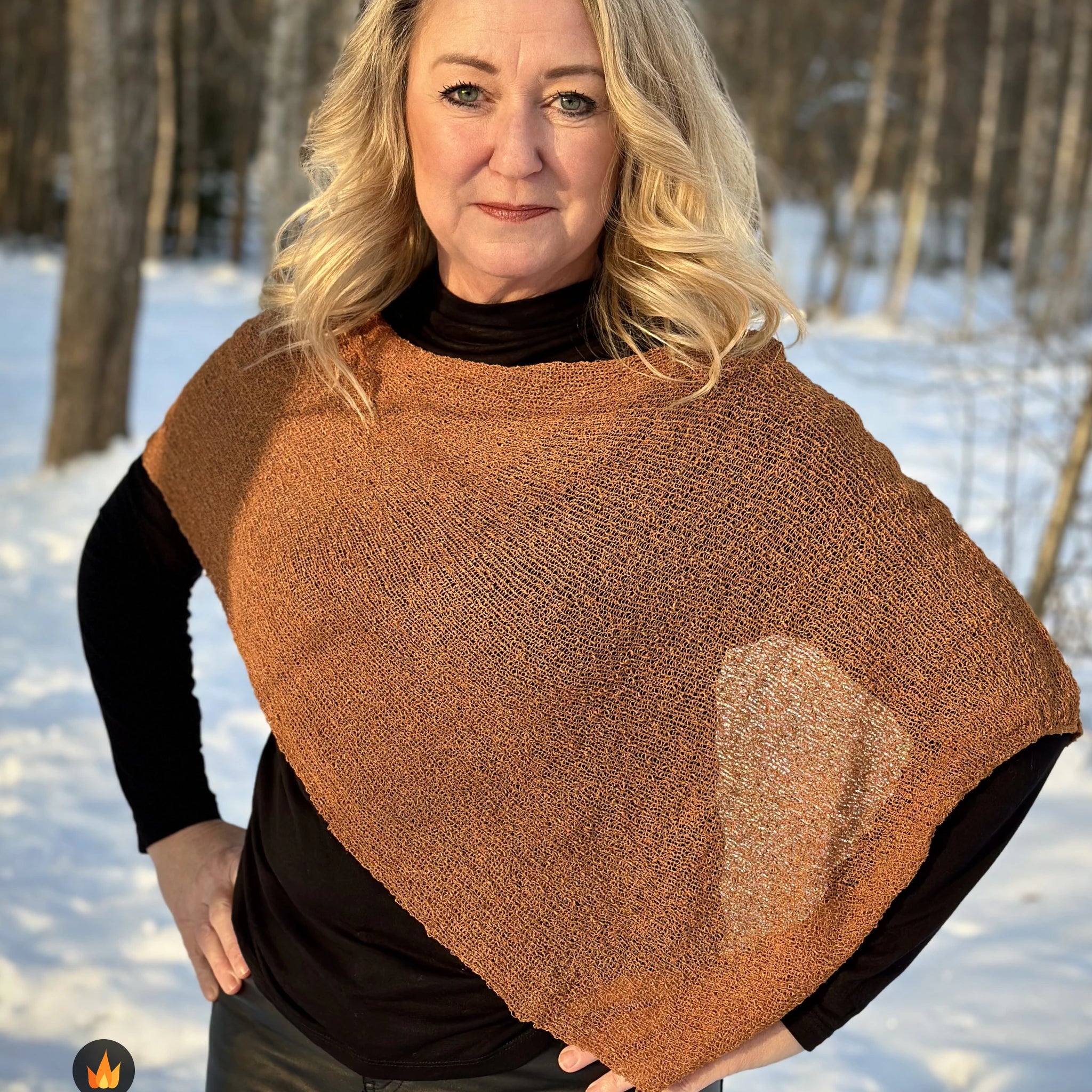 Kvinna bär Ella Cape i färg 009 Mocka, en varm färg i mockabrun med kopparglöd, finstickad cape som draperas mjukt över axlarna, fotad i motljus bland snöiga träd. Ulla Jacobsson Kläder.