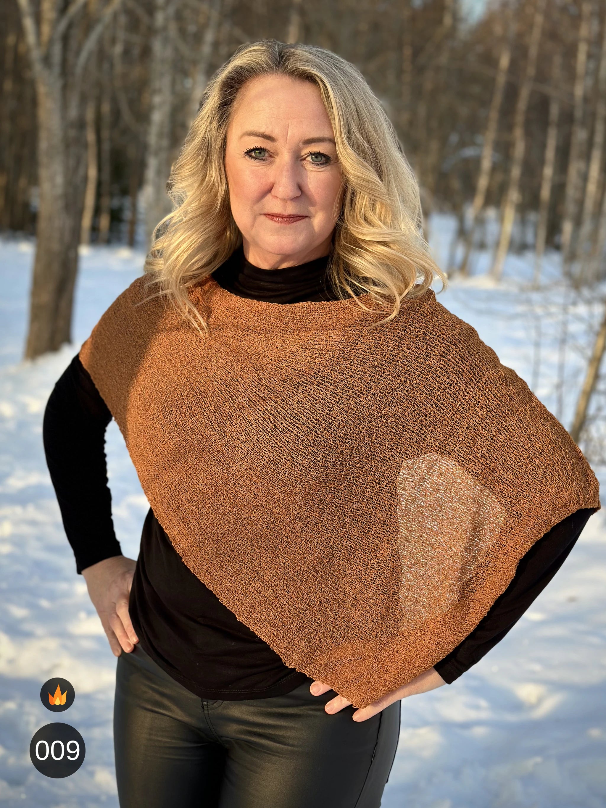 Kvinna bär Ella Cape i färg 009 Mocka, en varm färg i mockabrun med kopparglöd, finstickad cape som draperas mjukt över axlarna, fotad i motljus bland snöiga träd. Ulla Jacobsson Kläder.