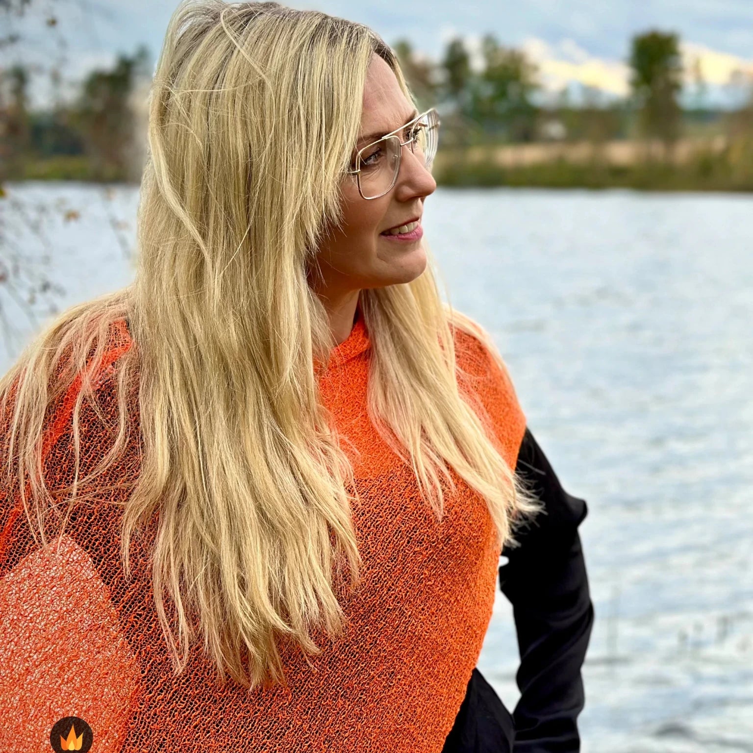 Kvinna bär Ella Cape i färg 011 Apelsin, en varm färg i klar apelsinorange, finstickad cape som draperas mjukt över axlarna, fotad vid en sjö en höstdag. Ulla Jacobsson Kläder.