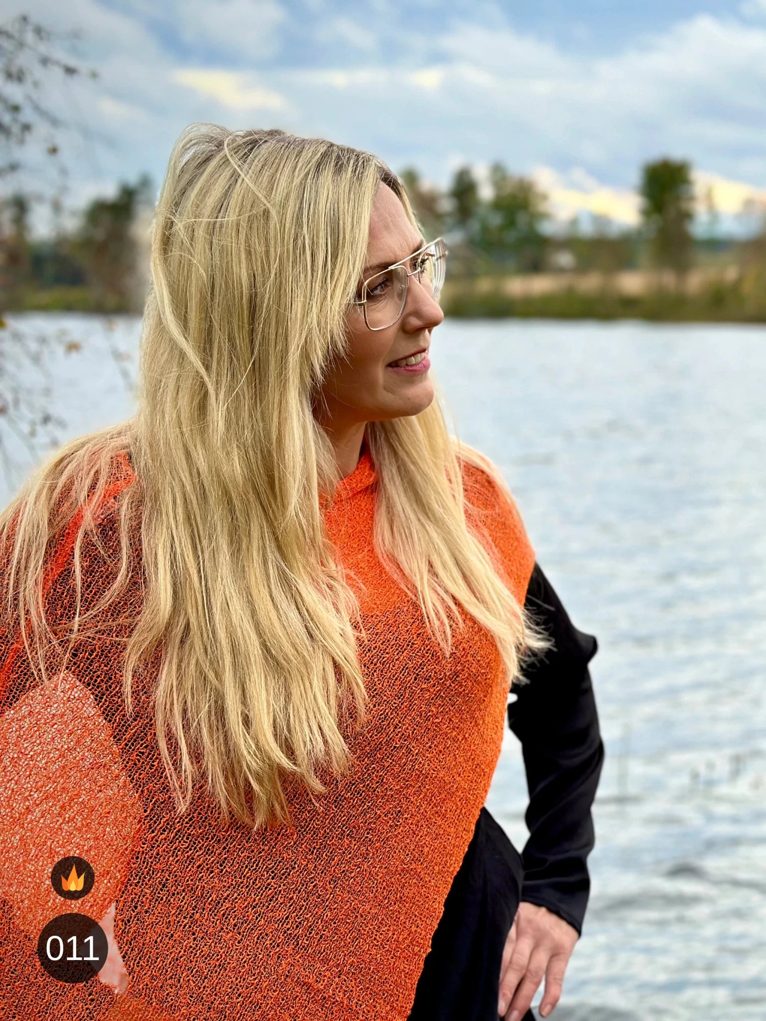 Kvinna bär Ella Cape i färg 011 Apelsin, en varm färg i klar apelsinorange, finstickad cape som draperas mjukt över axlarna, fotad vid en sjö en höstdag. Ulla Jacobsson Kläder.