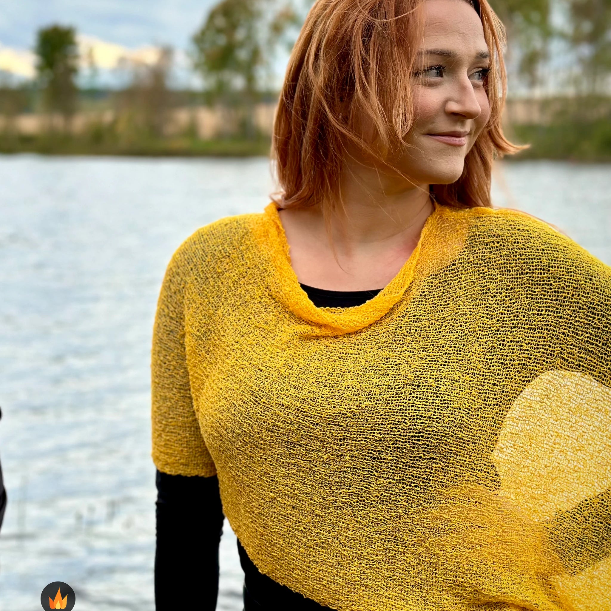Kvinna bär Ella Cape i färg 013 Saffran, en varm färg i djup saffransgul med gyllene glöd, finstickad cape som draperas mjukt över axlarna, fotad vid en sjö en höstdag. Ulla Jacobsson Kläder.