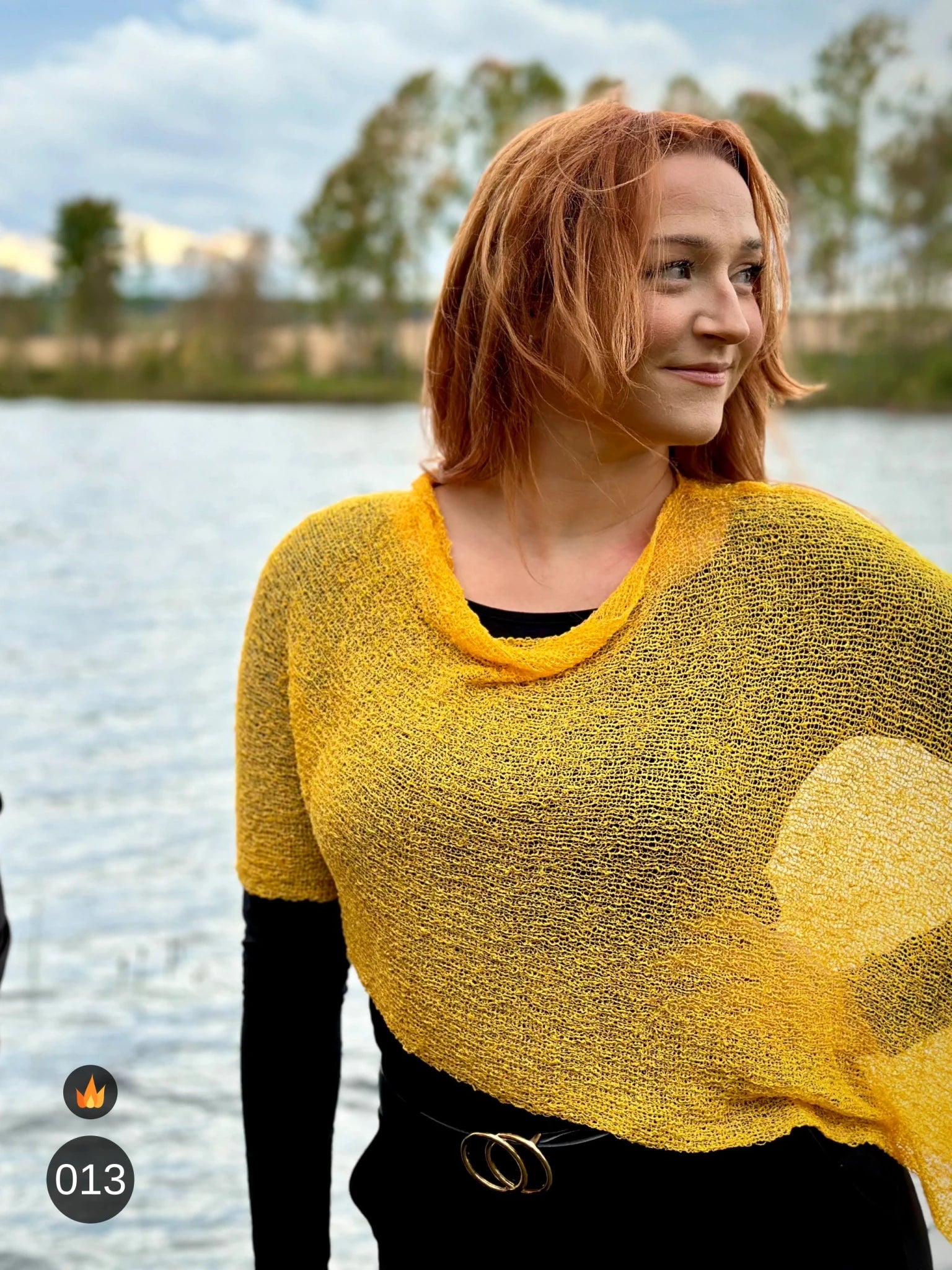 Kvinna bär Ella Cape i färg 013 Saffran, en varm färg i djup saffransgul med gyllene glöd, finstickad cape som draperas mjukt över axlarna, fotad vid en sjö en höstdag. Ulla Jacobsson Kläder.