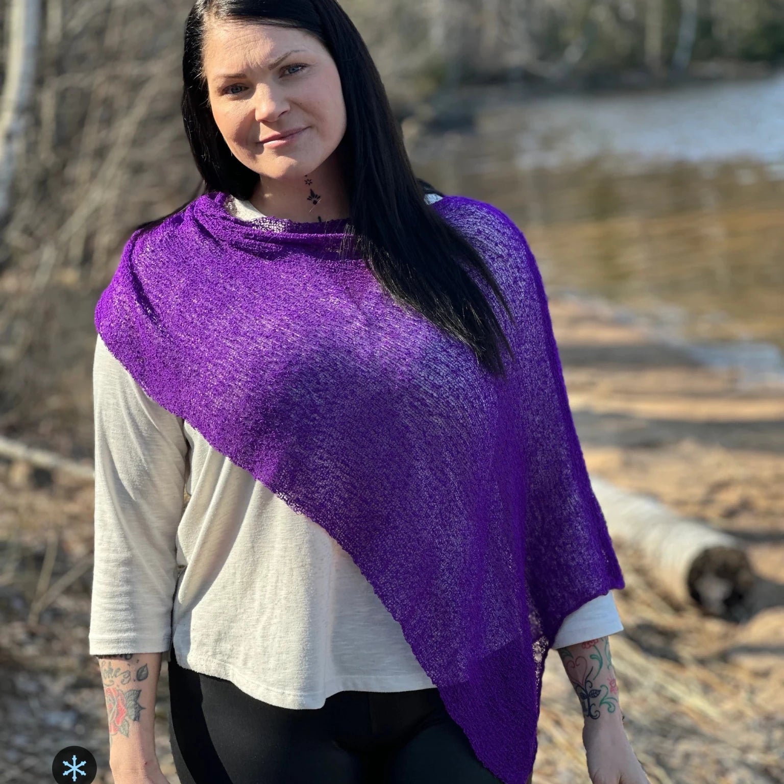 Kvinna bär Ella Cape i färg 014 Mörklila, en kall färg i djup violett lila, finstickad cape som draperas diagonalt över ena skuldran, fotad vid en å i vårljus. Ulla Jacobsson Kläder.