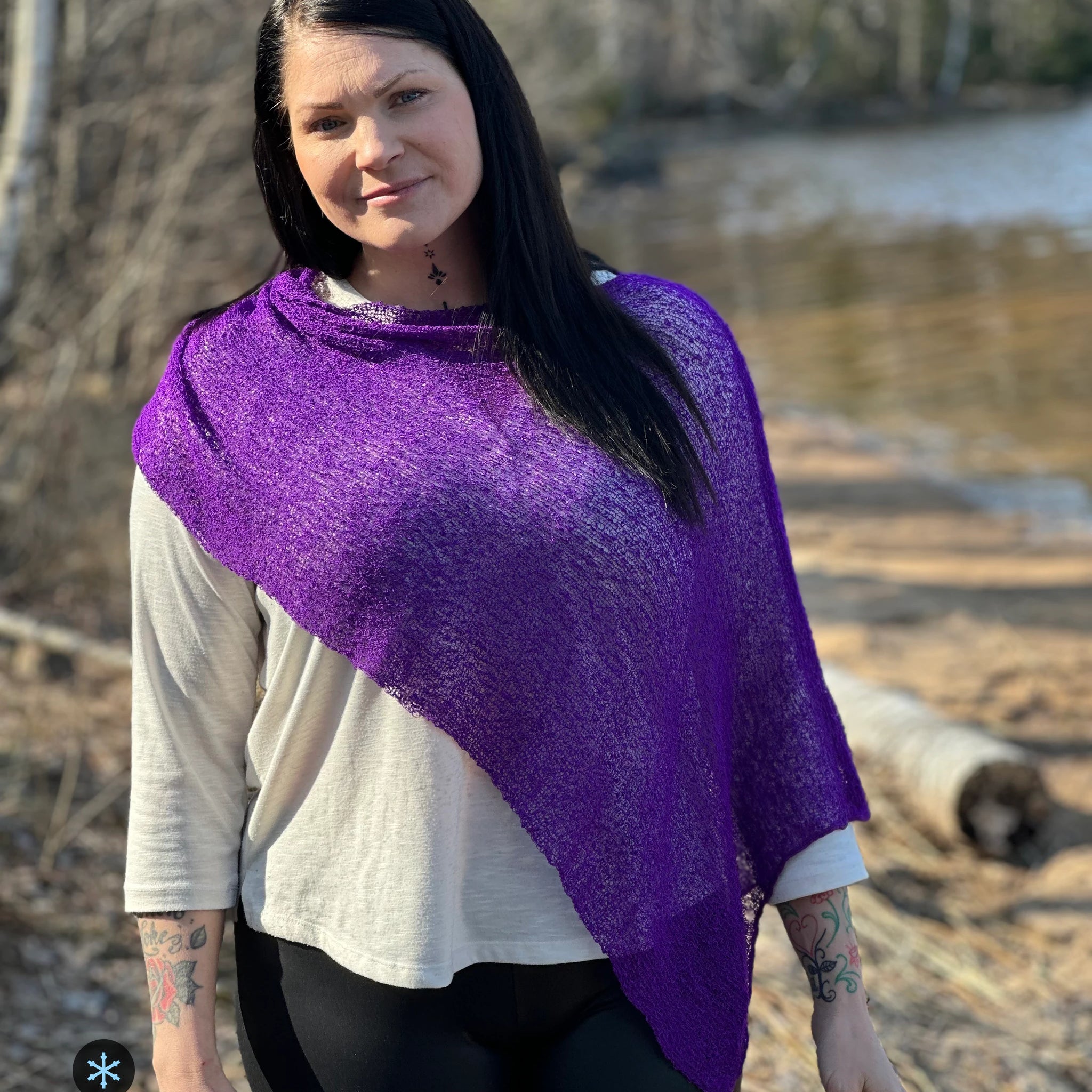 Kvinna bär Ella Cape i färg 014 Mörklila, en kall färg i djup violett lila, finstickad cape som draperas diagonalt över ena skuldran, fotad vid en å i vårljus. Ulla Jacobsson Kläder.